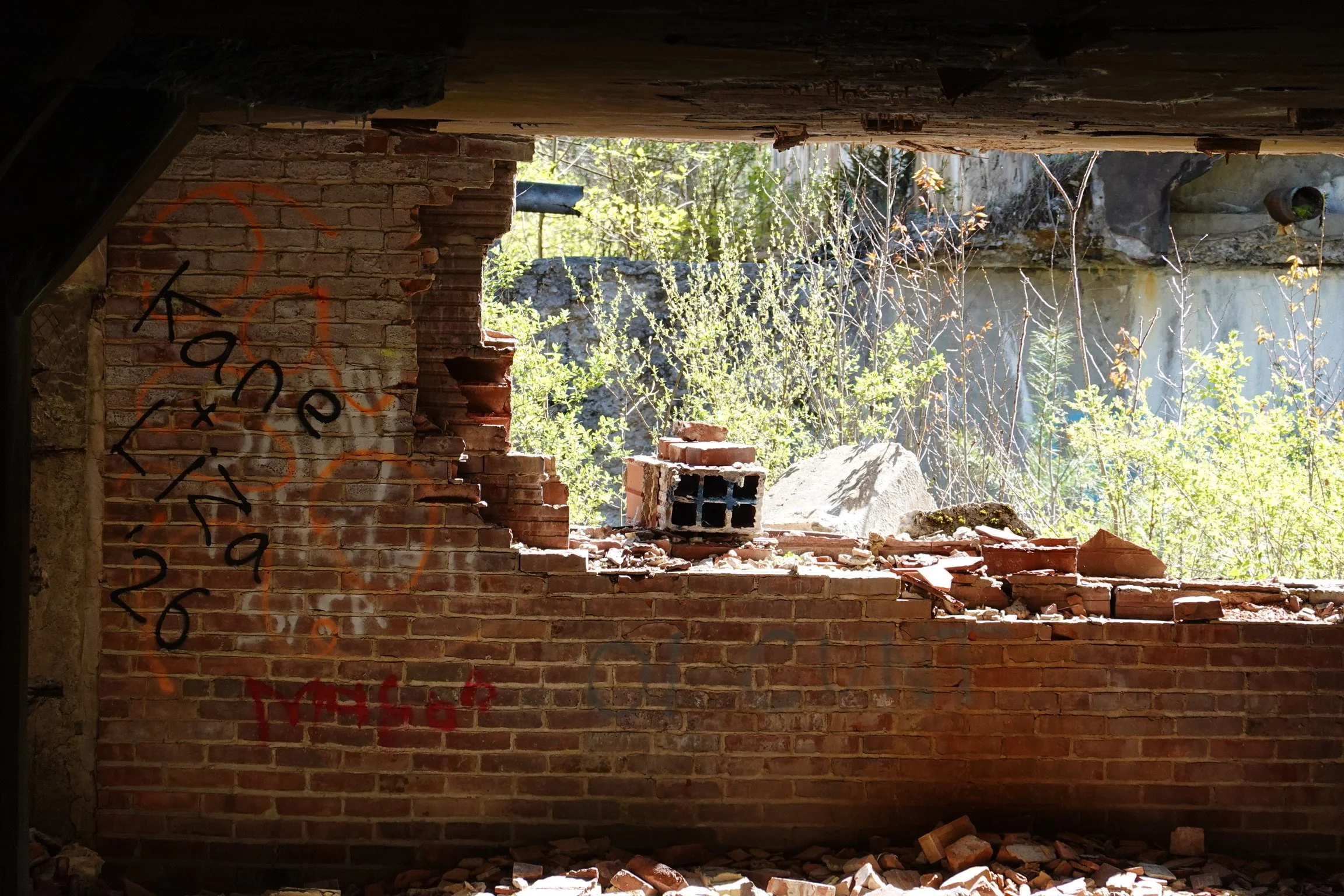 A partially demolished brick wall with graffiti and rubble inside a building, with trees and sunlight visible through the opening.
