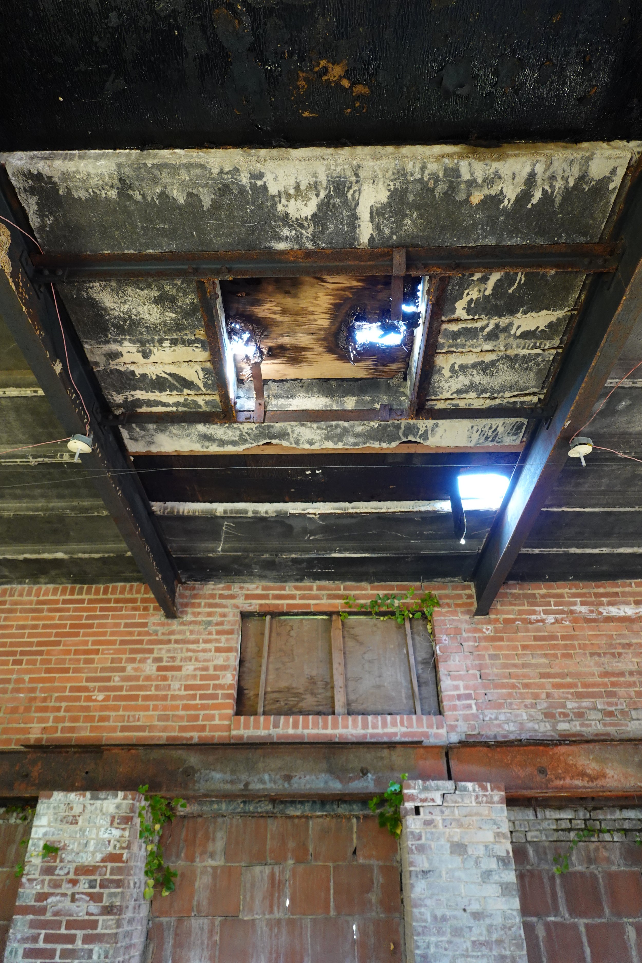 The image shows a ceiling structure with missing panels, revealing a rusted metal frame and damaged, burned wood behind it. The ceiling appears to be in a state of disrepair, with some boards charred and others broken, and there are small patches of 