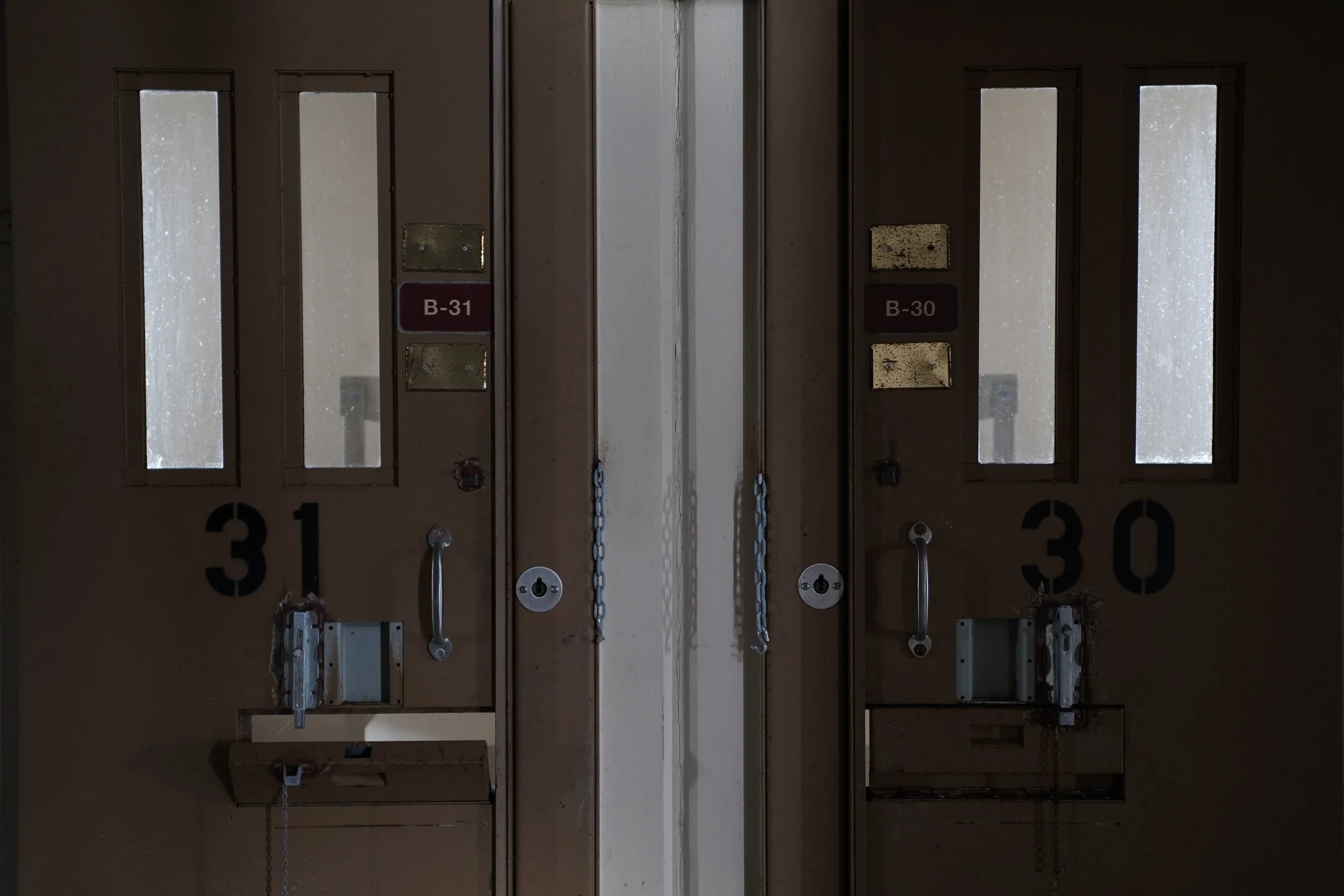 Image of closed metal lockers with labels B-31 and B-30. The lockers have small, dirty glass windows, locks, and chains securing the doors. The lockers are numbered 31 and 30, with visible handles.
