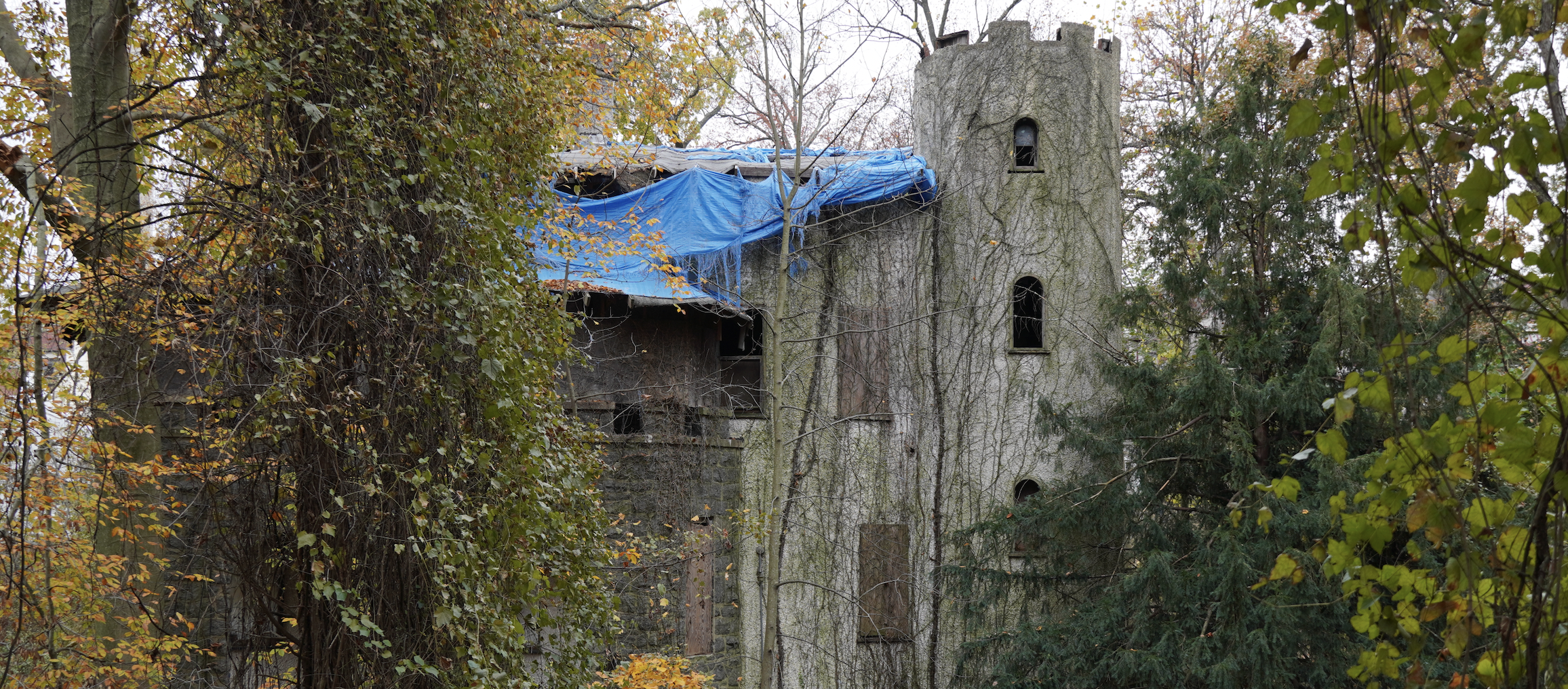 An old, abandoned stone castle tower and building covered in vines, surrounded by trees with autumn leaves, some trees with green and yellow foliage and some with bare branches.