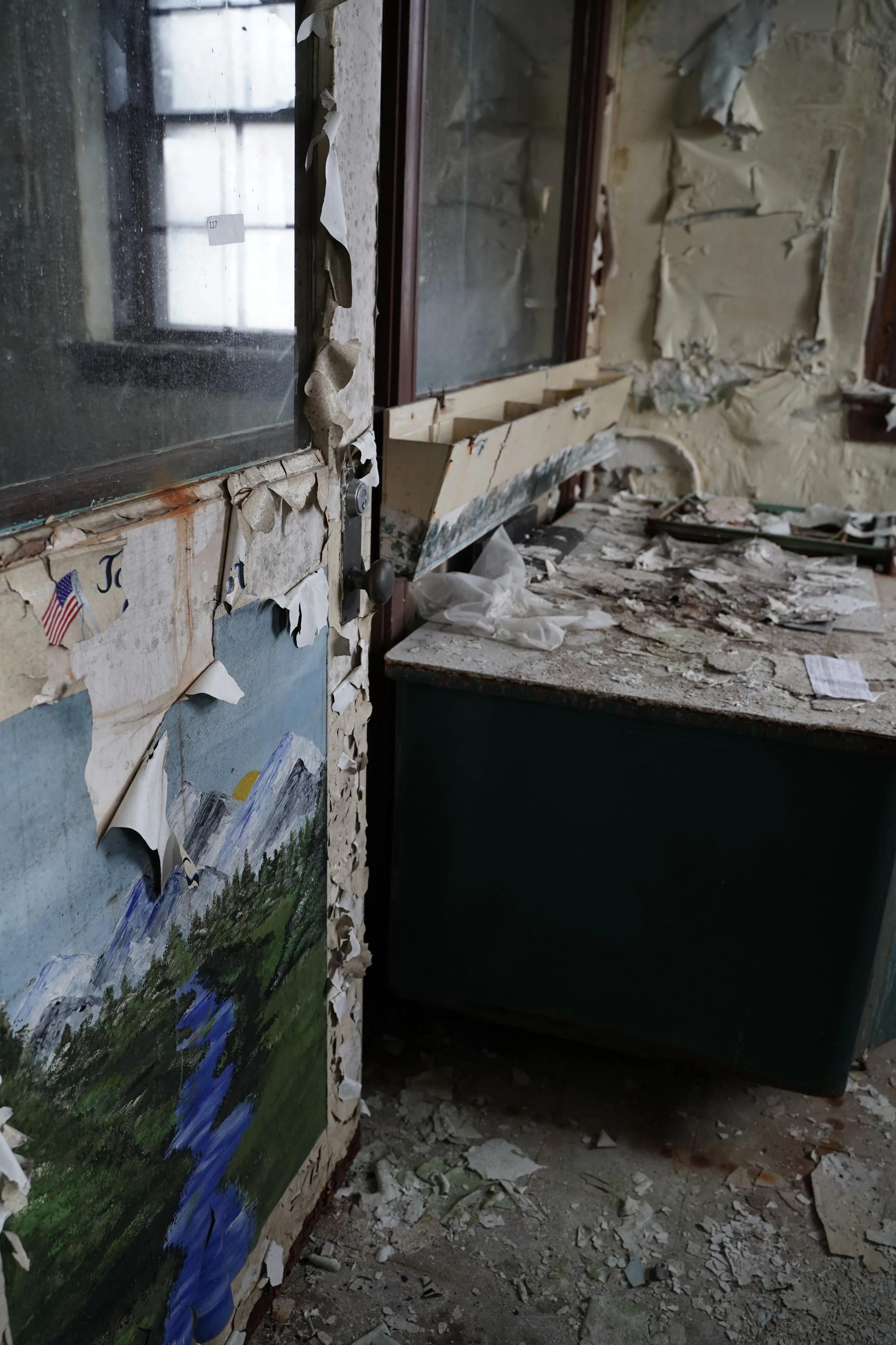 A damaged and peeling wall inside an abandoned building, with debris on the floor and a landscape painting leaning against the wall.