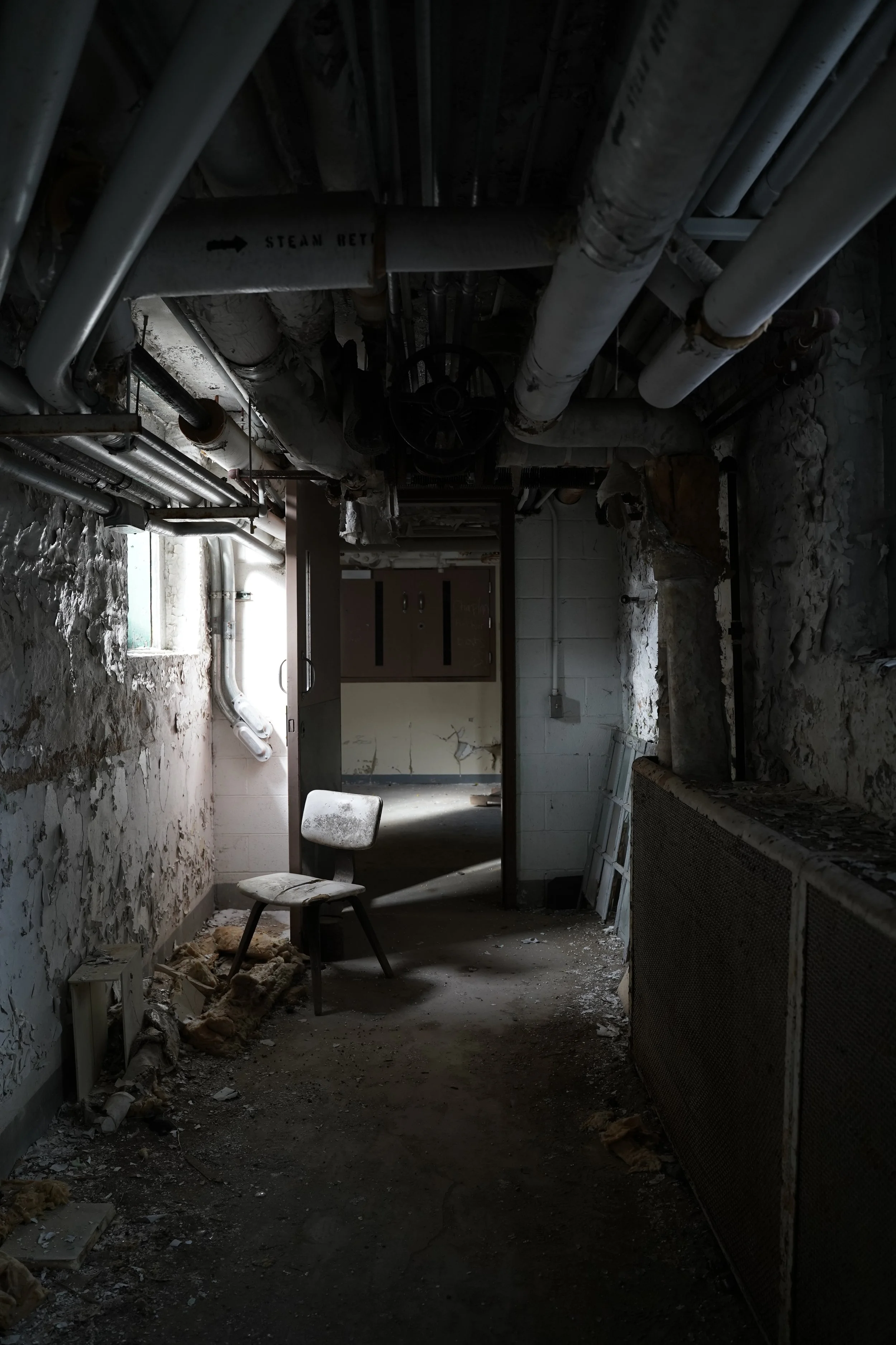 An abandoned and dusty basement room with peeling paint on the walls, exposed pipes on the ceiling, a metal chair with a torn cushion, debris on the floor, and a small window letting in some light.