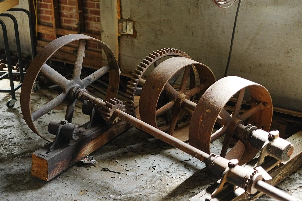An old mechanical device with large rusty gears and wheels, set on a wooden base on a concrete floor in a workshop or garage.