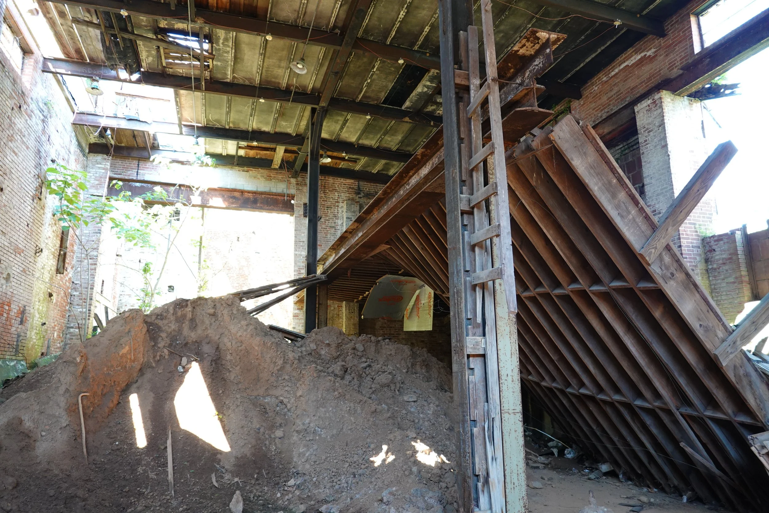 Construction site inside an old brick building with exposed structural elements, wooden stairs, and a pile of dirt in the foreground.