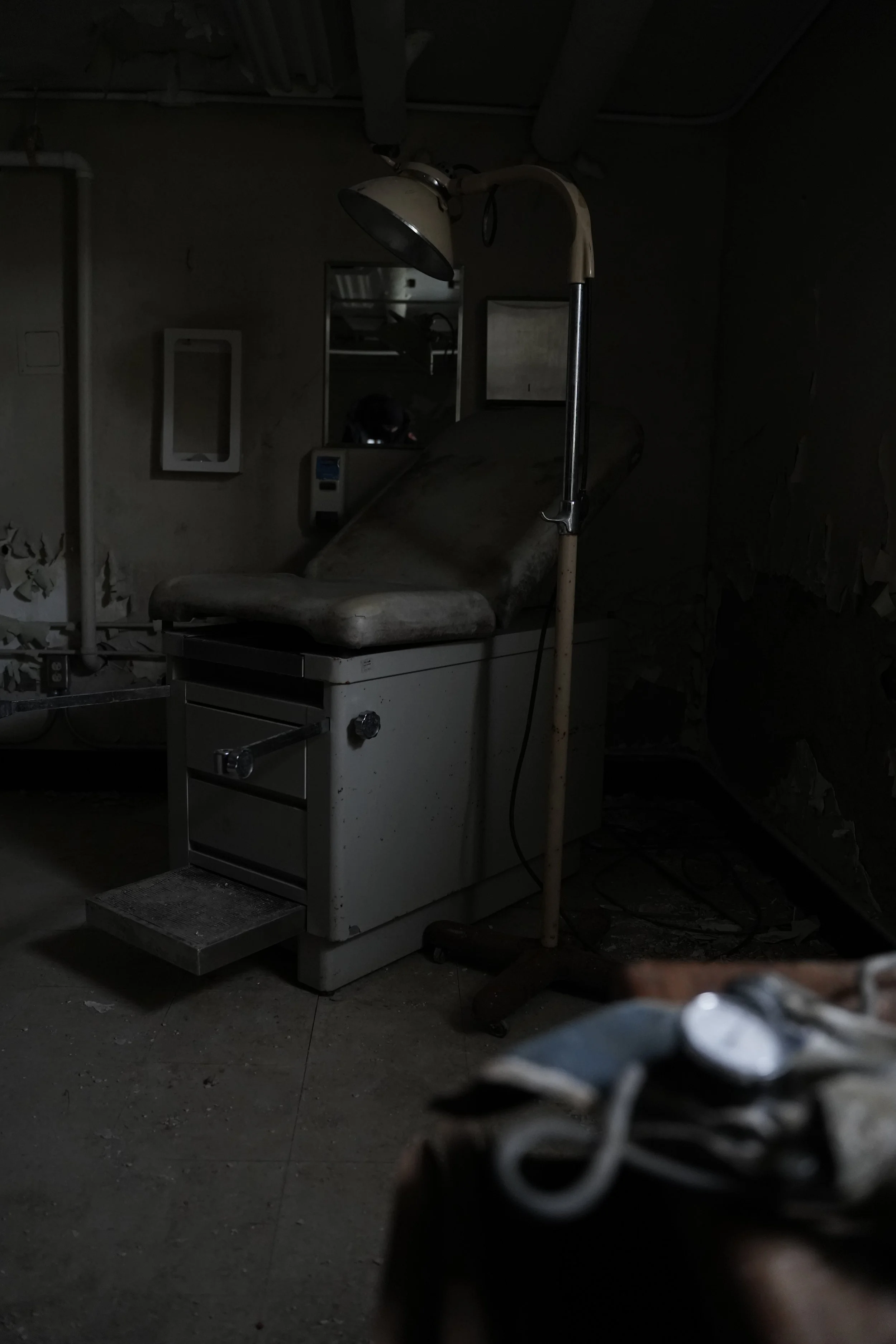 Dark, abandoned hospital room with a dusty, old medical examination table, a floor lamp, and peeling wallpaper.