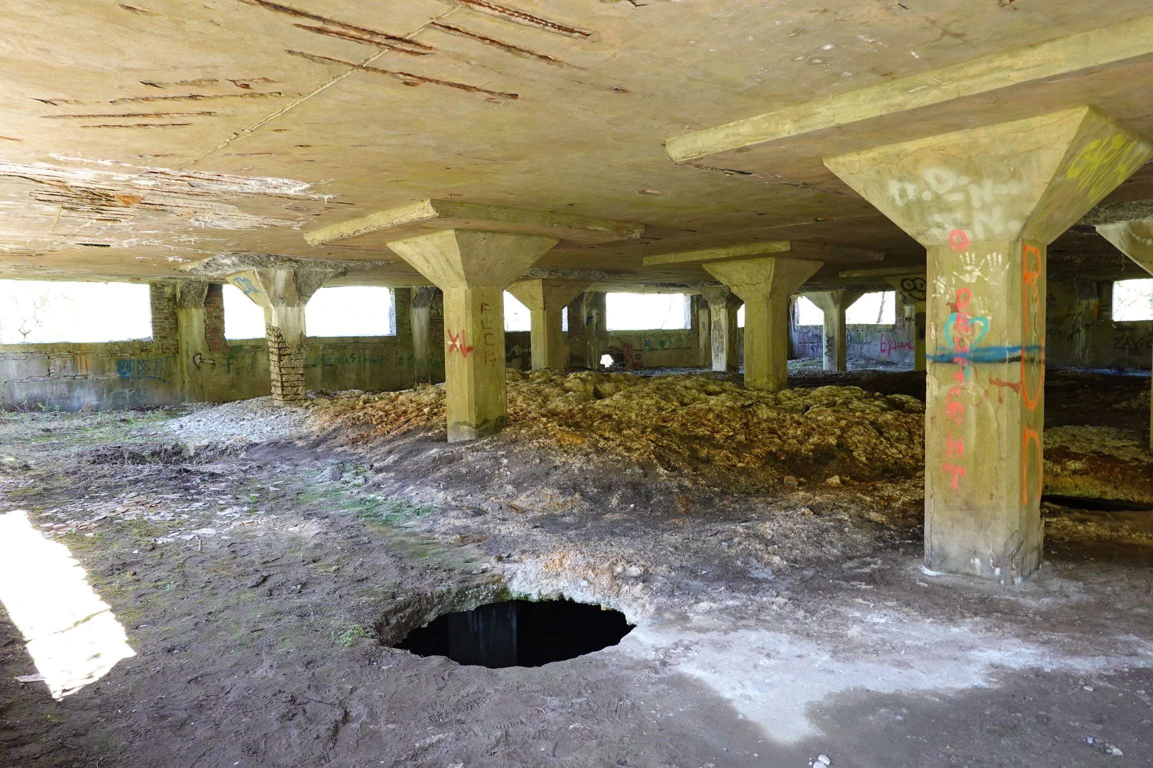 An abandoned building with graffiti on the walls and support columns. The floor is uneven with dirt and debris, and there is a large hole in the ground.