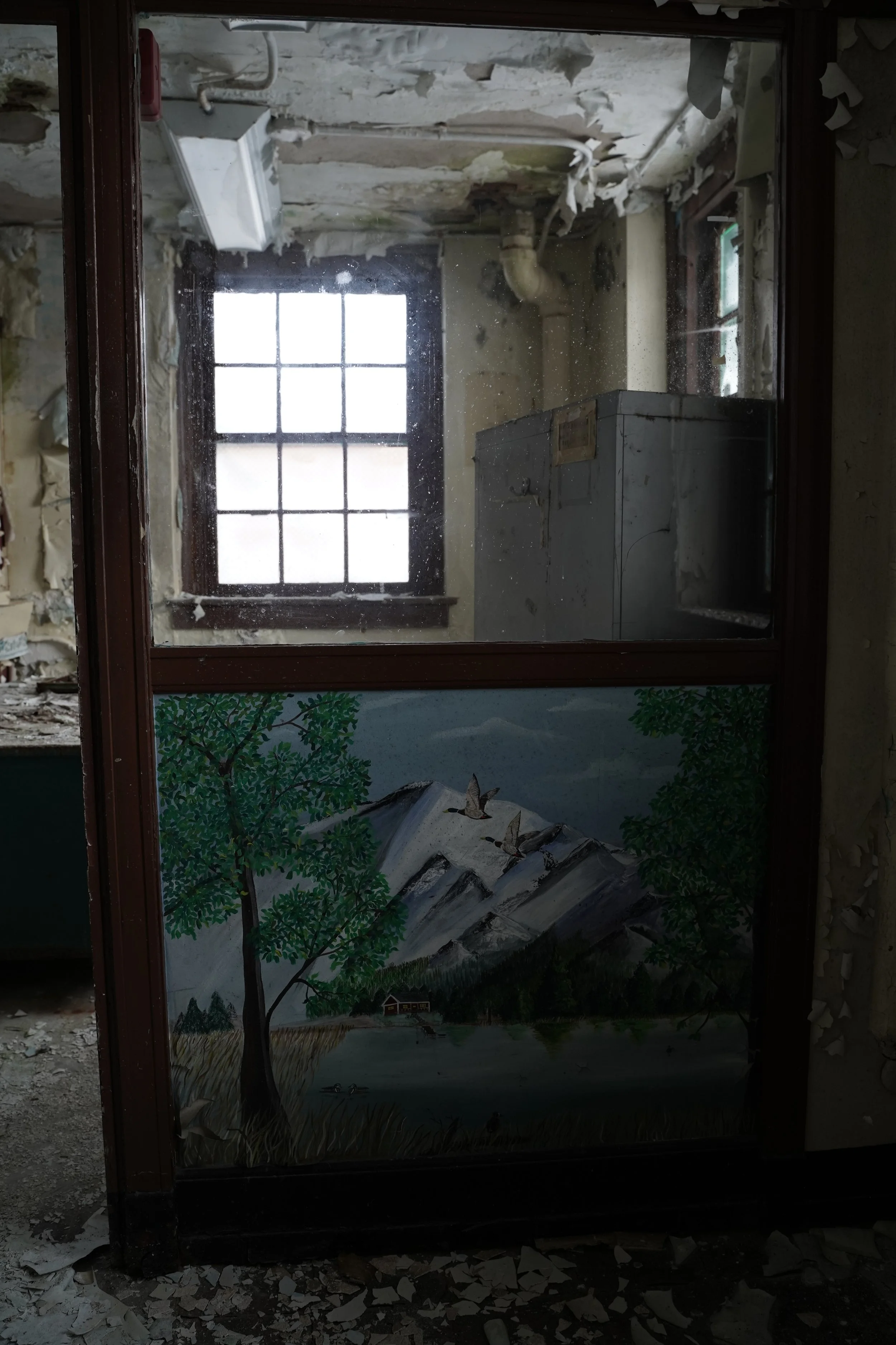 A broken mirror reflects a mural with trees, a house, a river, and a mountain landscape, with debris and rubble surrounding it.