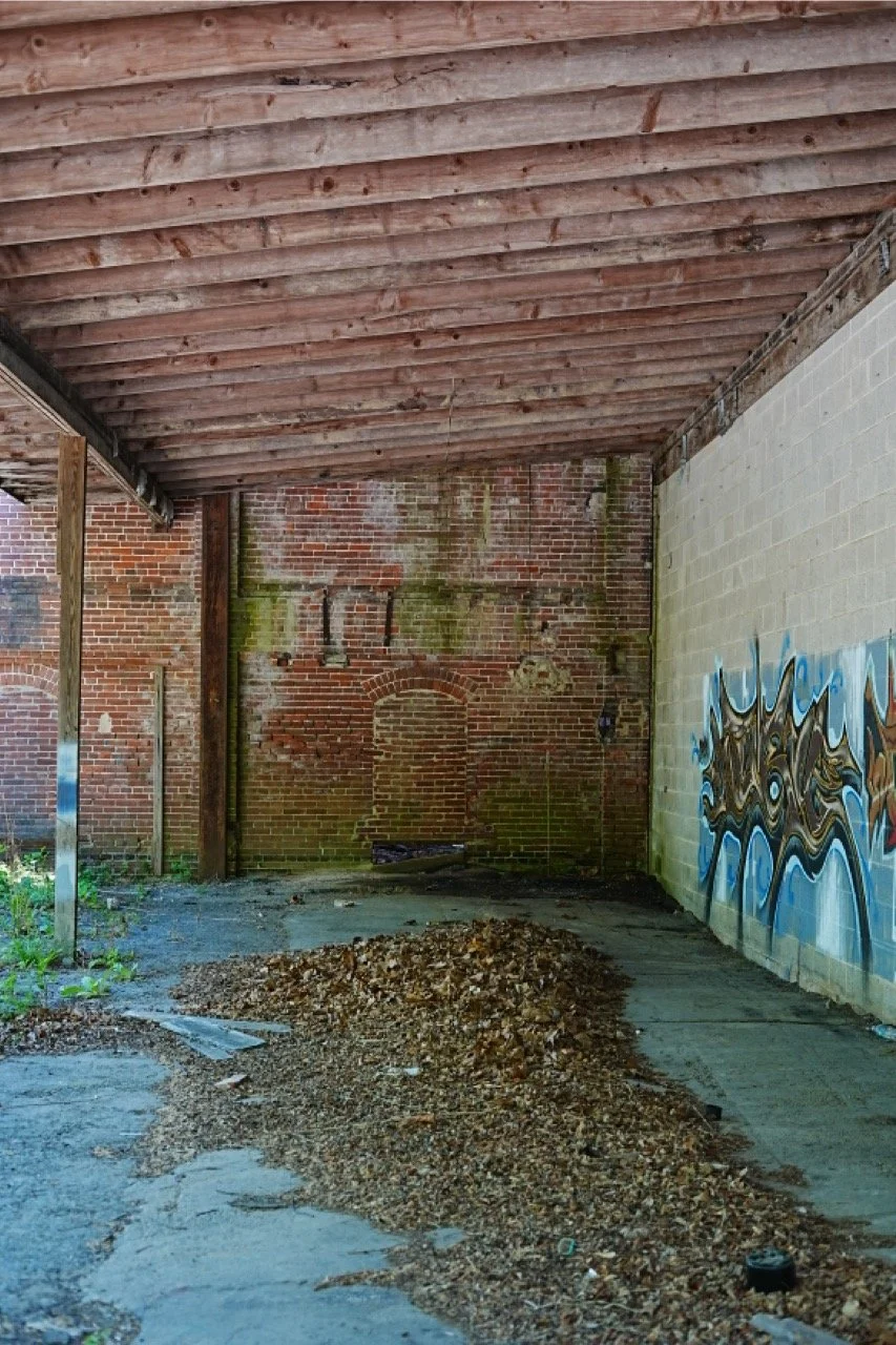 An abandoned outdoor space with a wooden roof and brick walls, some covered in moss and graffiti. The ground has a pile of leaves and debris, with cracked concrete and overgrown plants.
