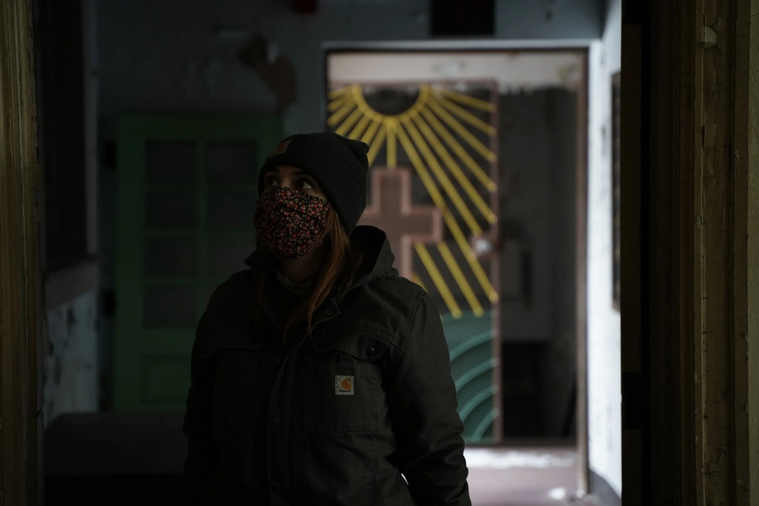 A person wearing a black beanie and a patterned face mask standing inside a structure, looking to the side, with a background featuring a yellow and black pathway pattern and a cross symbol.