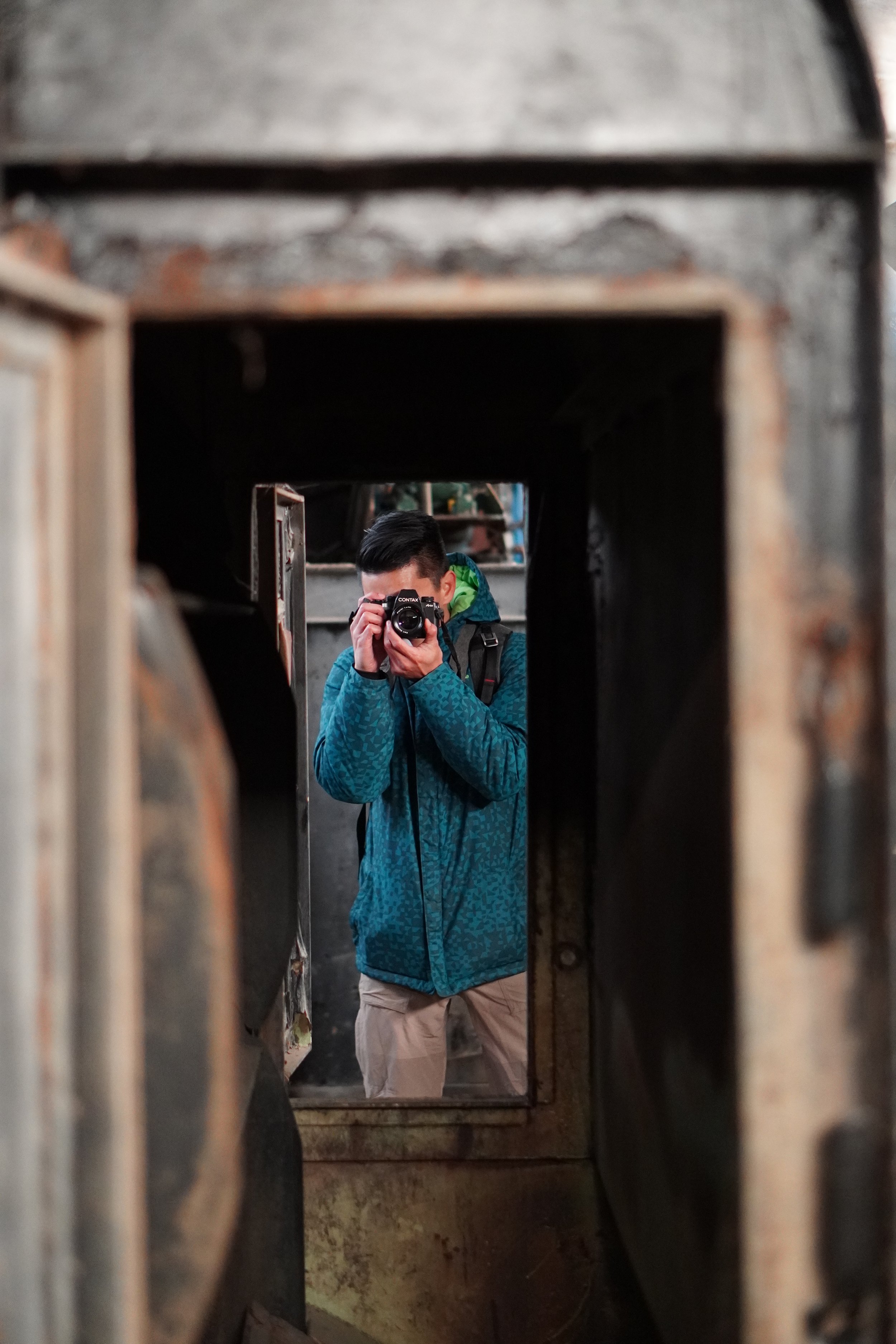 A man in a blue patterned jacket takes a photo of himself using a camera, seen through a narrow, rusted metal opening.