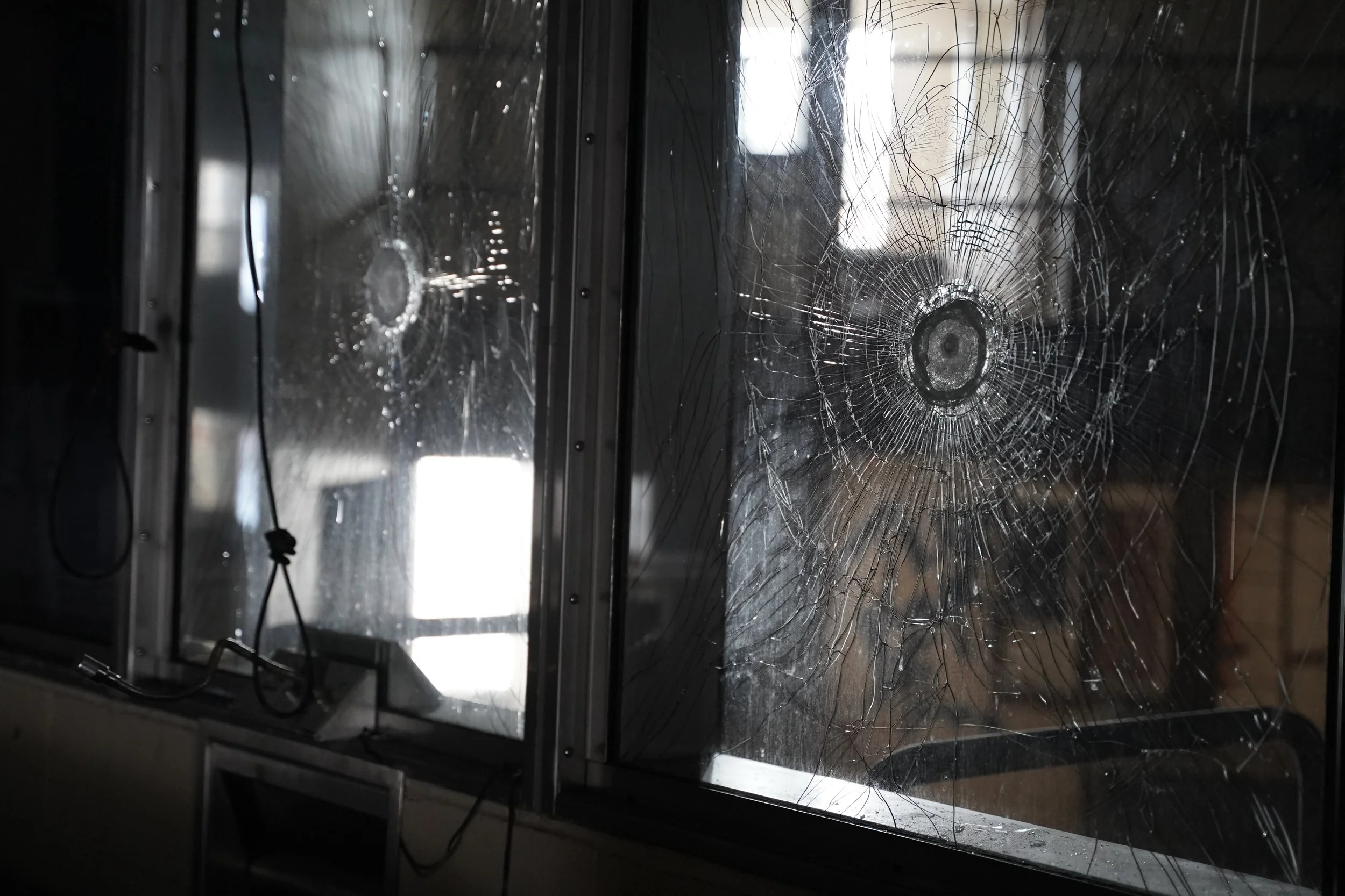 Shattered glass window with prominent cracks and spiderweb pattern, reflected in a metallic surface, with some wires visible.