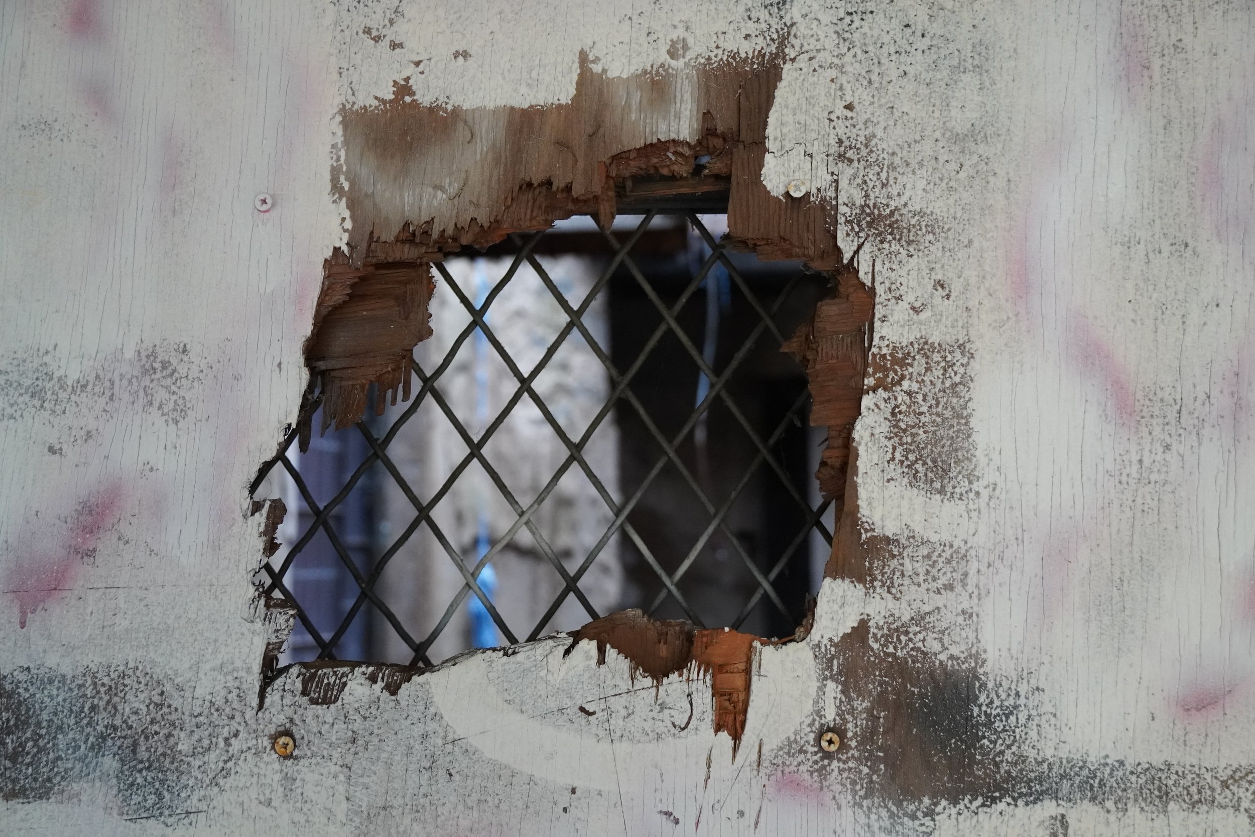 A damaged drywall with a hole revealing a metal wire mesh behind it, held in place by two screws.