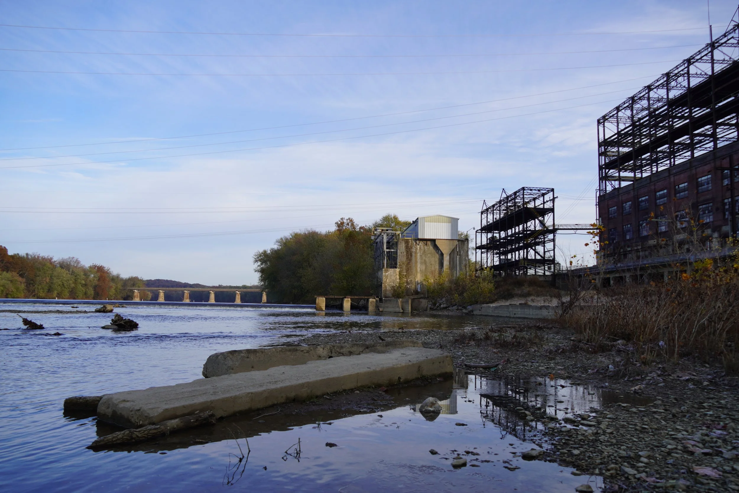 River with rocks along the shore, industrial buildings with metal frameworks, power lines, and trees in the background under a partly cloudy sky.