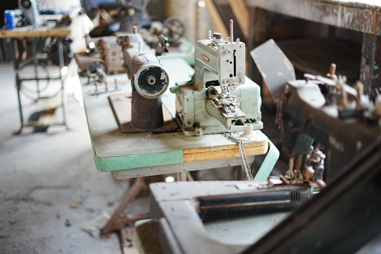 An old, rusted sewing machine on a worn green and beige workbench in a workshop surrounded by vintage mechanical parts and tools.