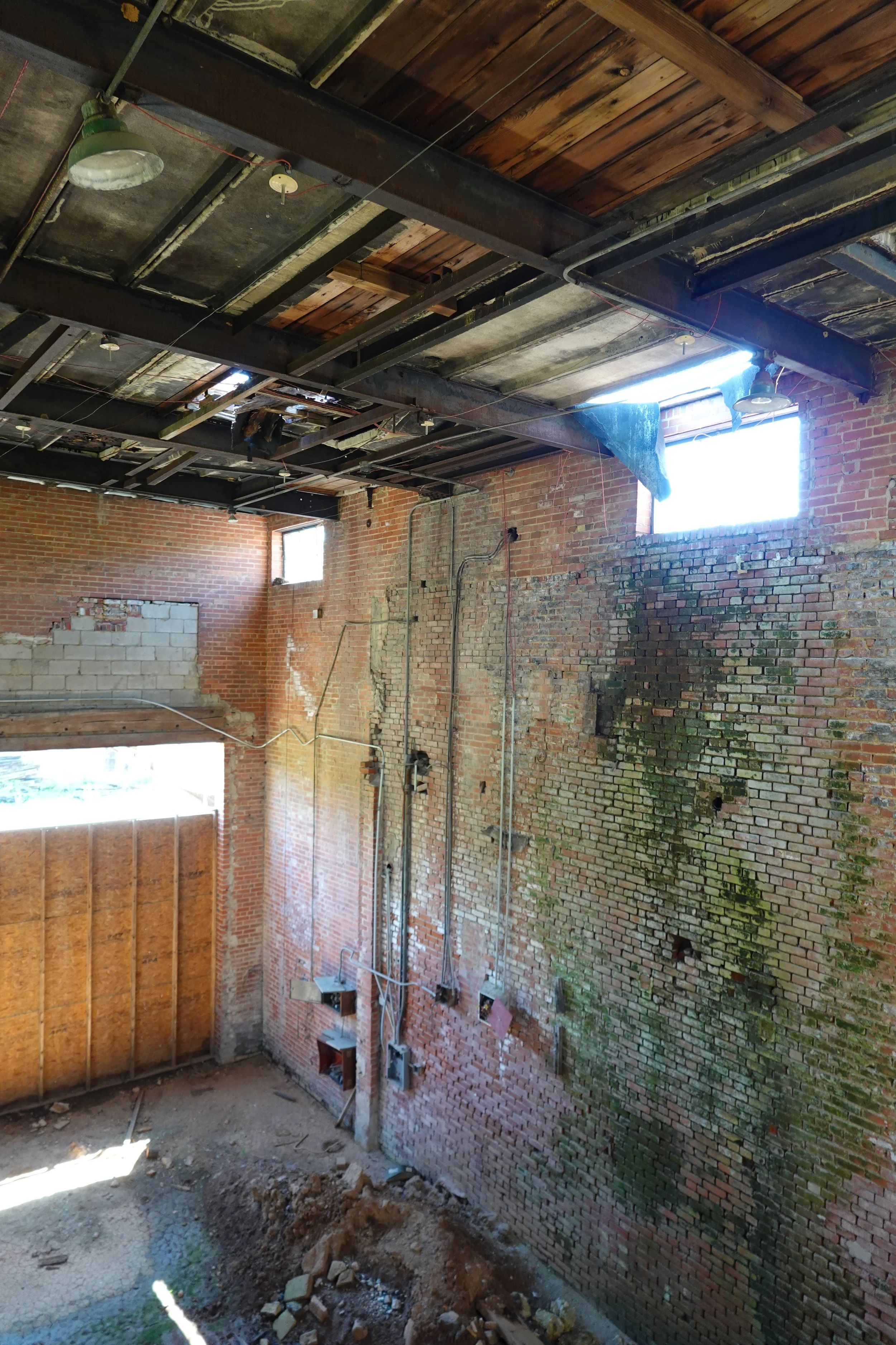 Interior view of a brick building under renovation, with exposed brick walls, a partially removed ceiling, and construction debris on the floor.