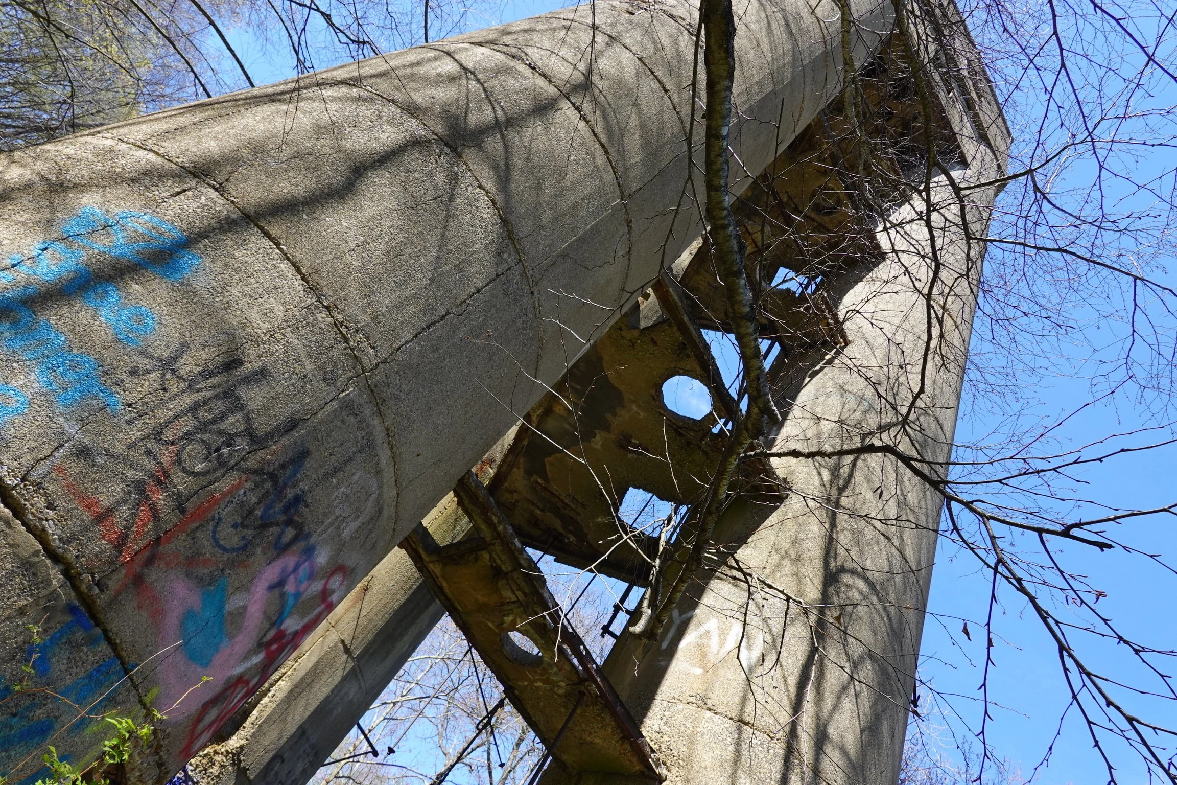 An abandoned concrete structure with arches and columns, covered in graffiti and surrounded by leafless trees under a clear blue sky.