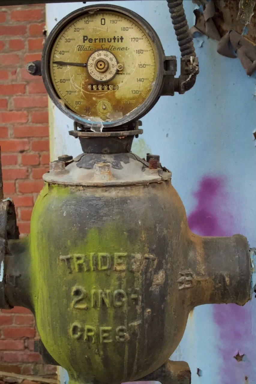 An antique water meter with a yellow face, featuring the brand name 'Permutit' and a dial indicating volume. The meter is attached to a rusty pipe and the object below is a dirty, old valve box with the inscription 'TRIDE 2 IN. GREG', with visible ru