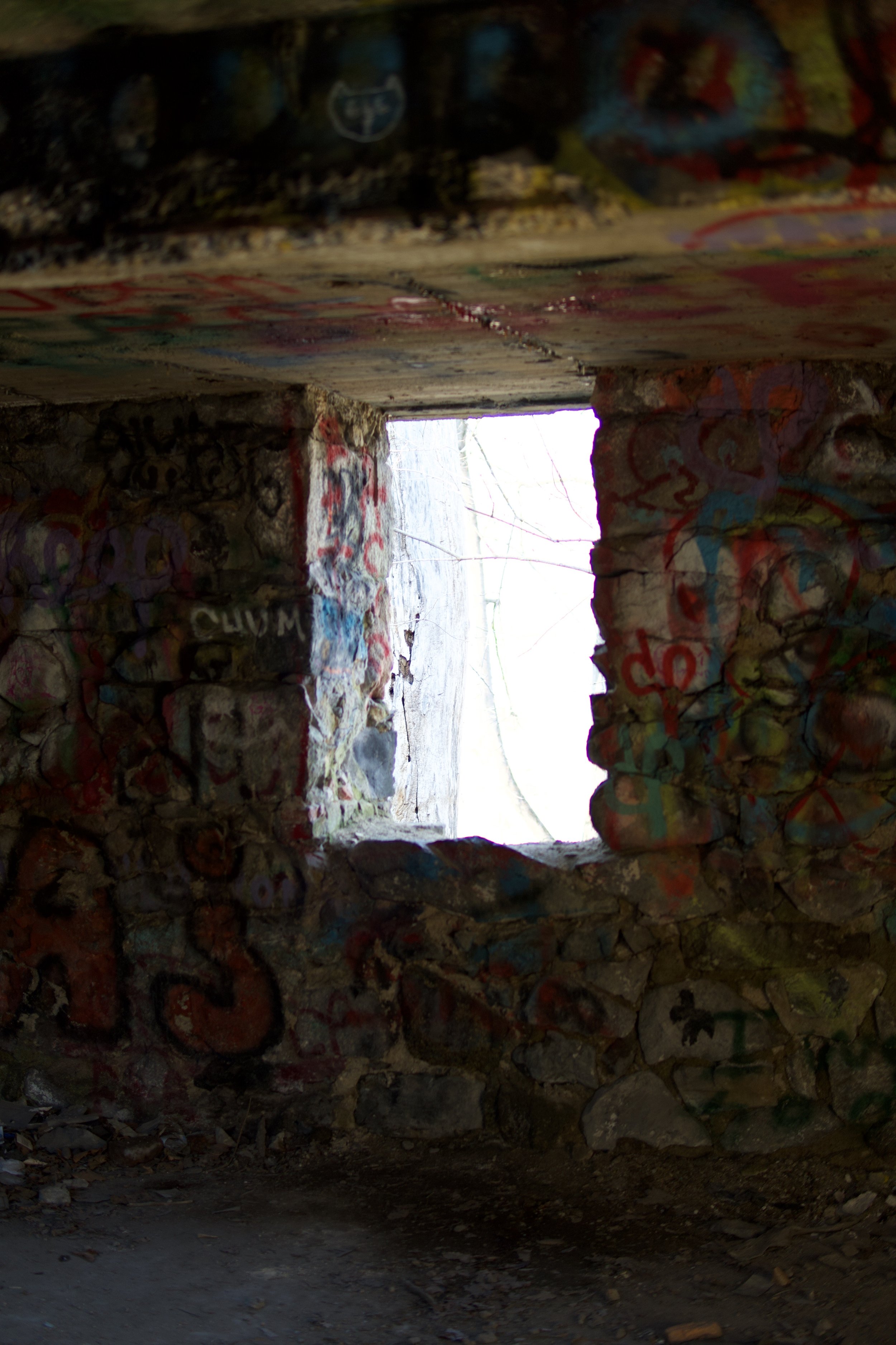 A photo taken from beneath a graffiti-covered bridge or structure during daytime, showing the rocky ground and the opening to the outside with visible trees and bright light.