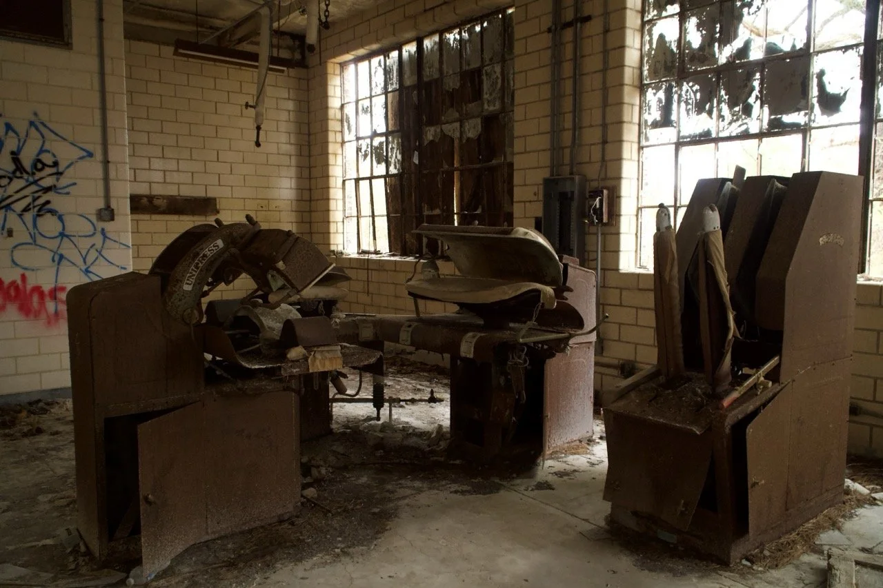 An abandoned, dusty office space with broken furniture and graffiti on the wall, illuminated by sunlight through cracked windows.