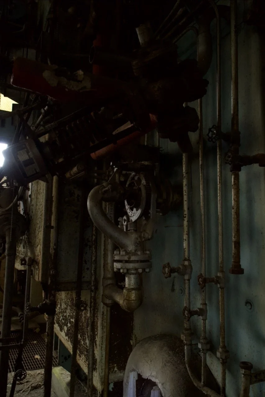 Rusty industrial pipes and machinery, including valves and a large curved pipe, mounted on a corroded metal wall.