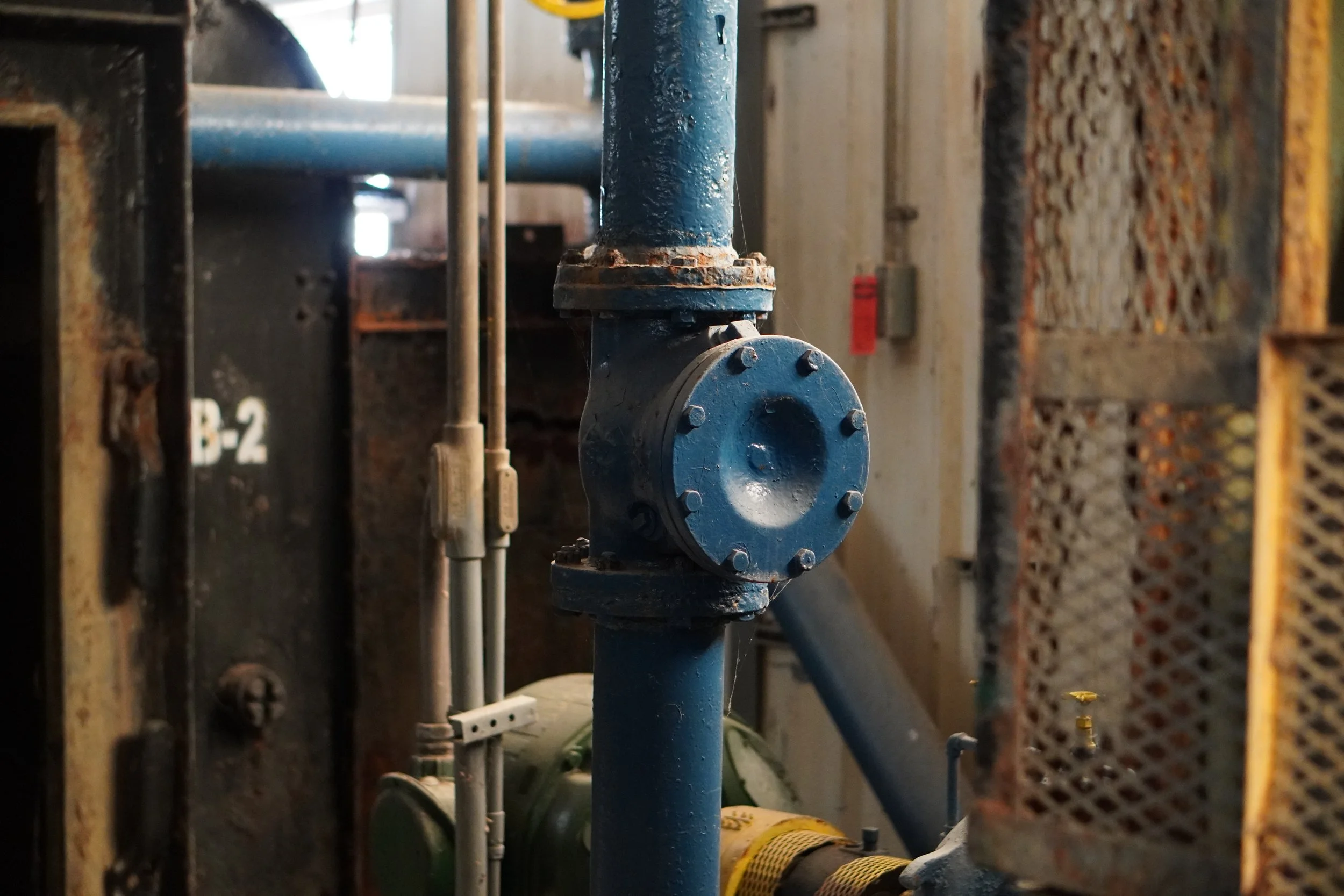 Close-up of an industrial water pipe system with a prominent blue pipe and rusted flange, surrounded by other pipes and machinery in a factory setting.