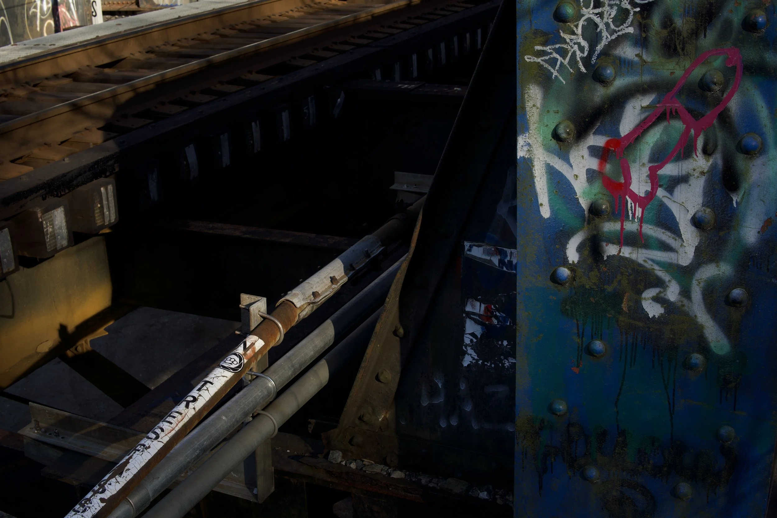 Close-up of graffiti art on a metal structure near railroad tracks, with pipes and bolts visible.