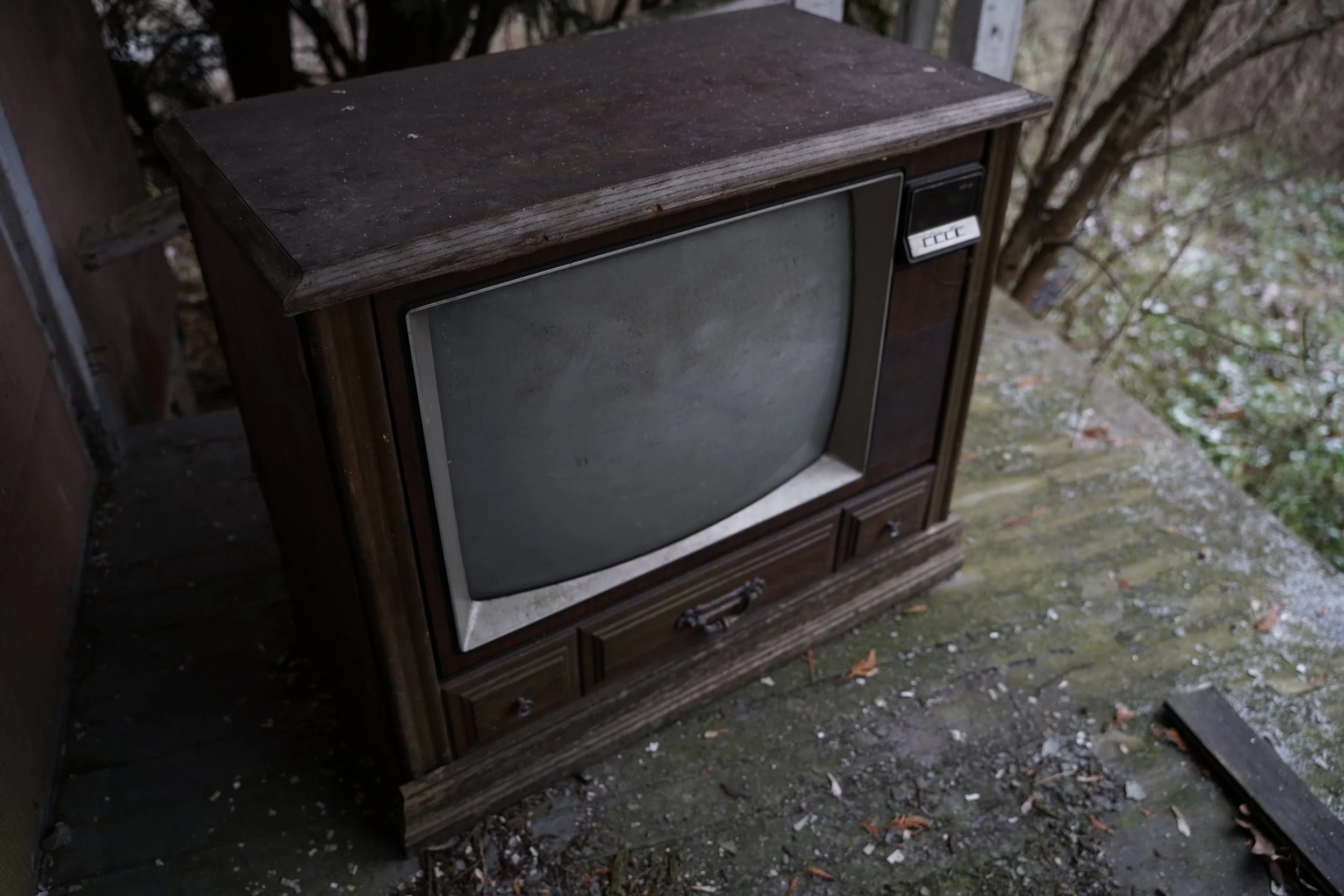 Old, weathered wooden cabinet with a glass door in an outdoor setting, surrounded by dirt and fallen leaves.