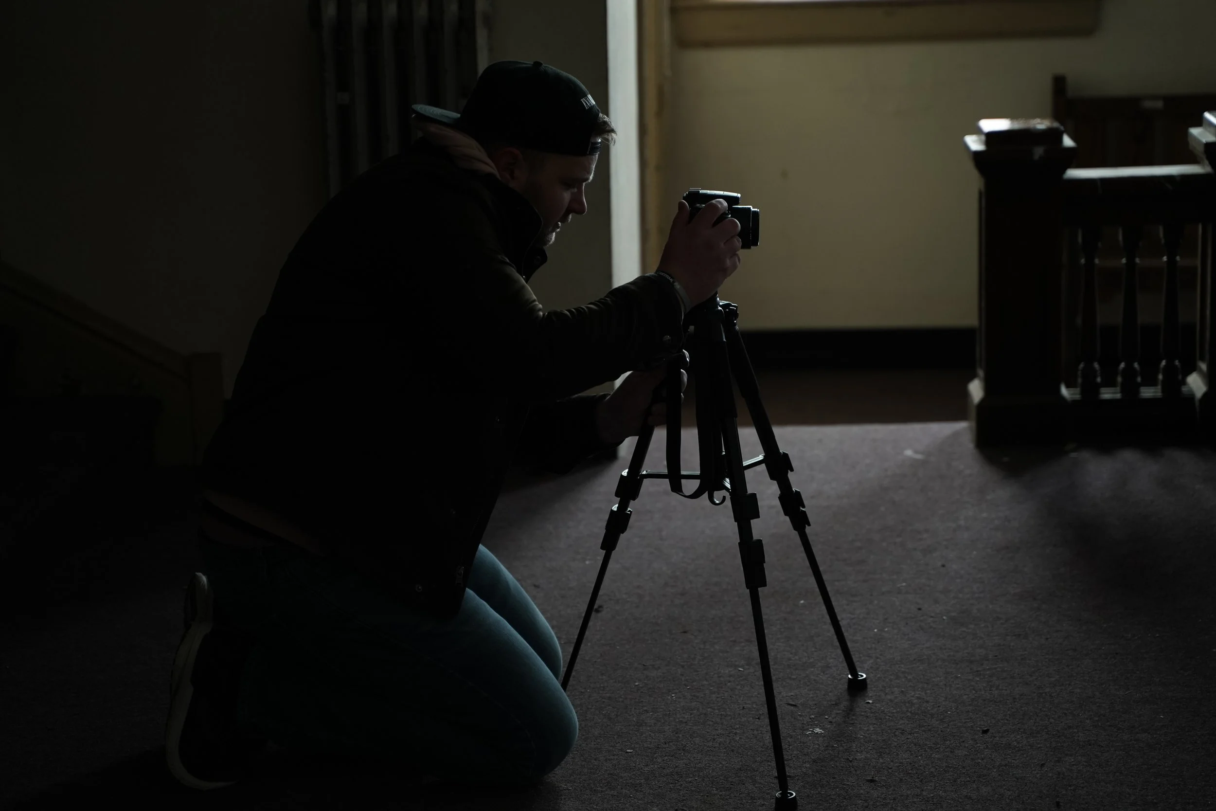 A person kneeling on the floor, adjusting a camera on a tripod in a dimly lit room near a window.