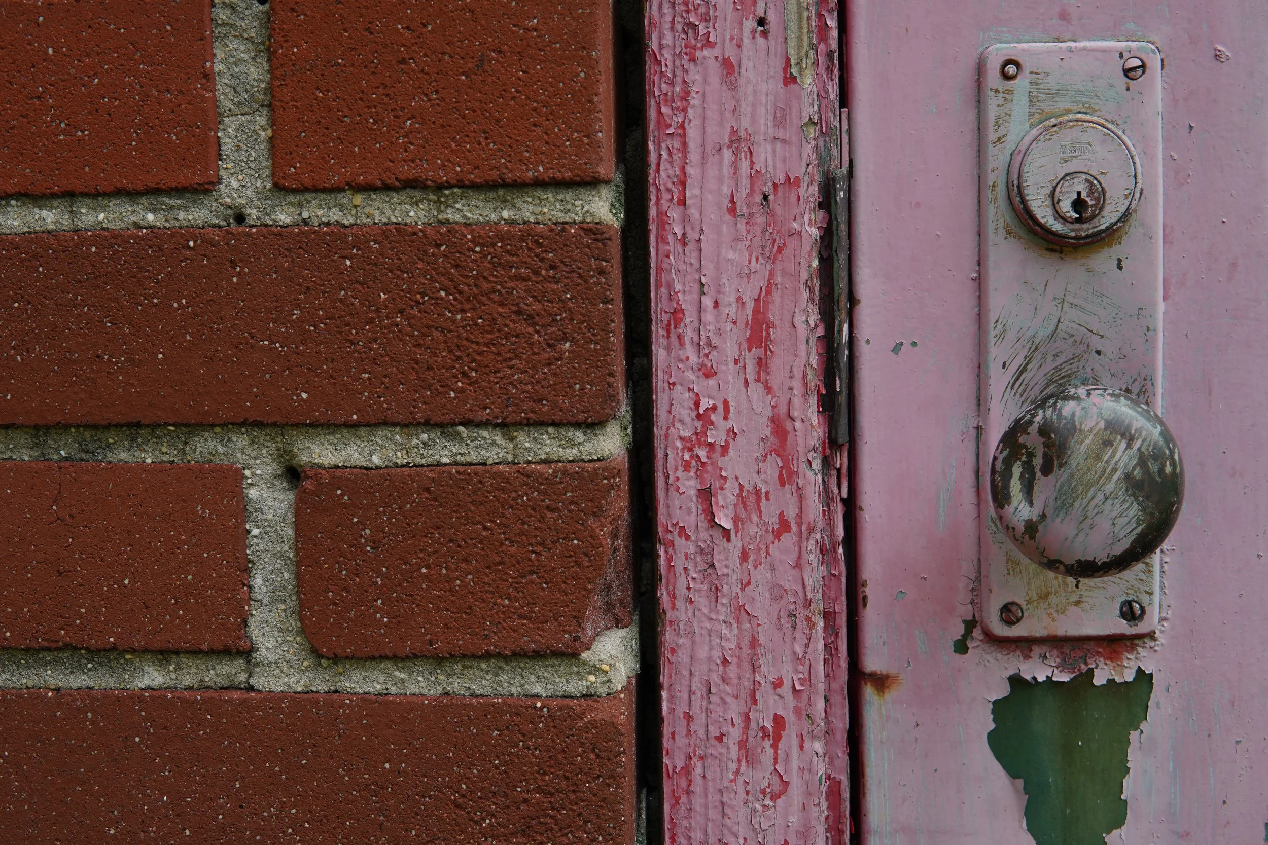 Close-up of a brick wall next to a weathered pink wooden door with a rusty doorknob and lock.