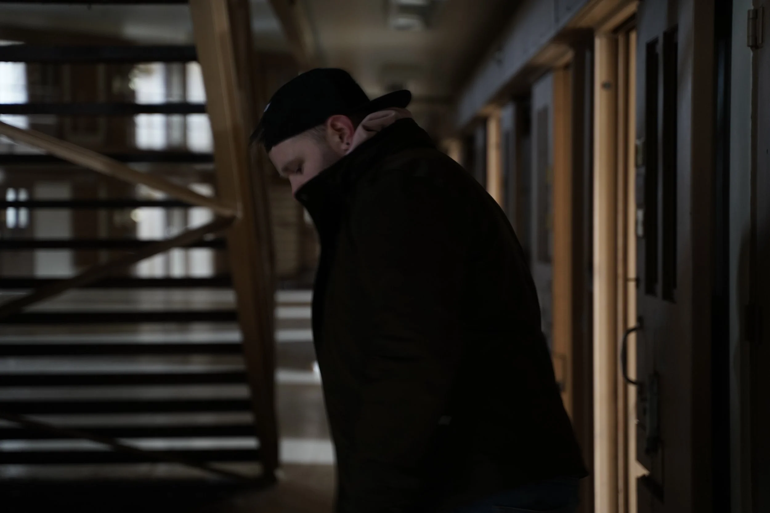A person wearing a black cap and dark clothing standing inside a house, looking down, with wooden stairs and framed pictures on the wall in the background.