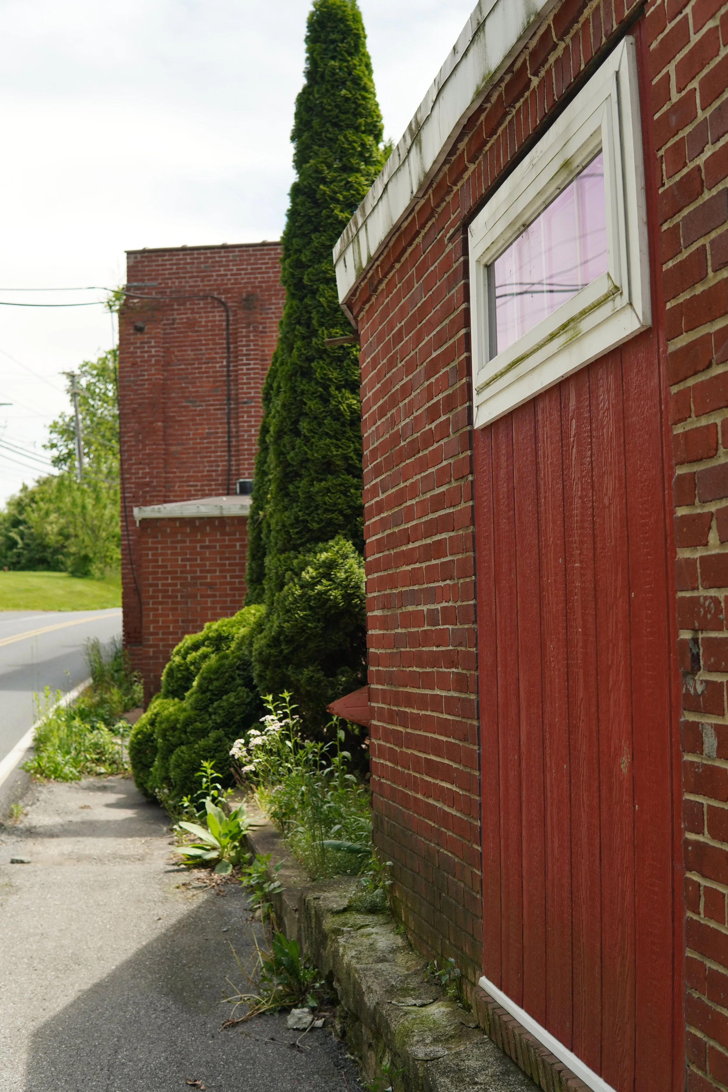 Close-up of a red brick building with a red wooden door, surrounded by green bushes and plants, and a narrow sidewalk.