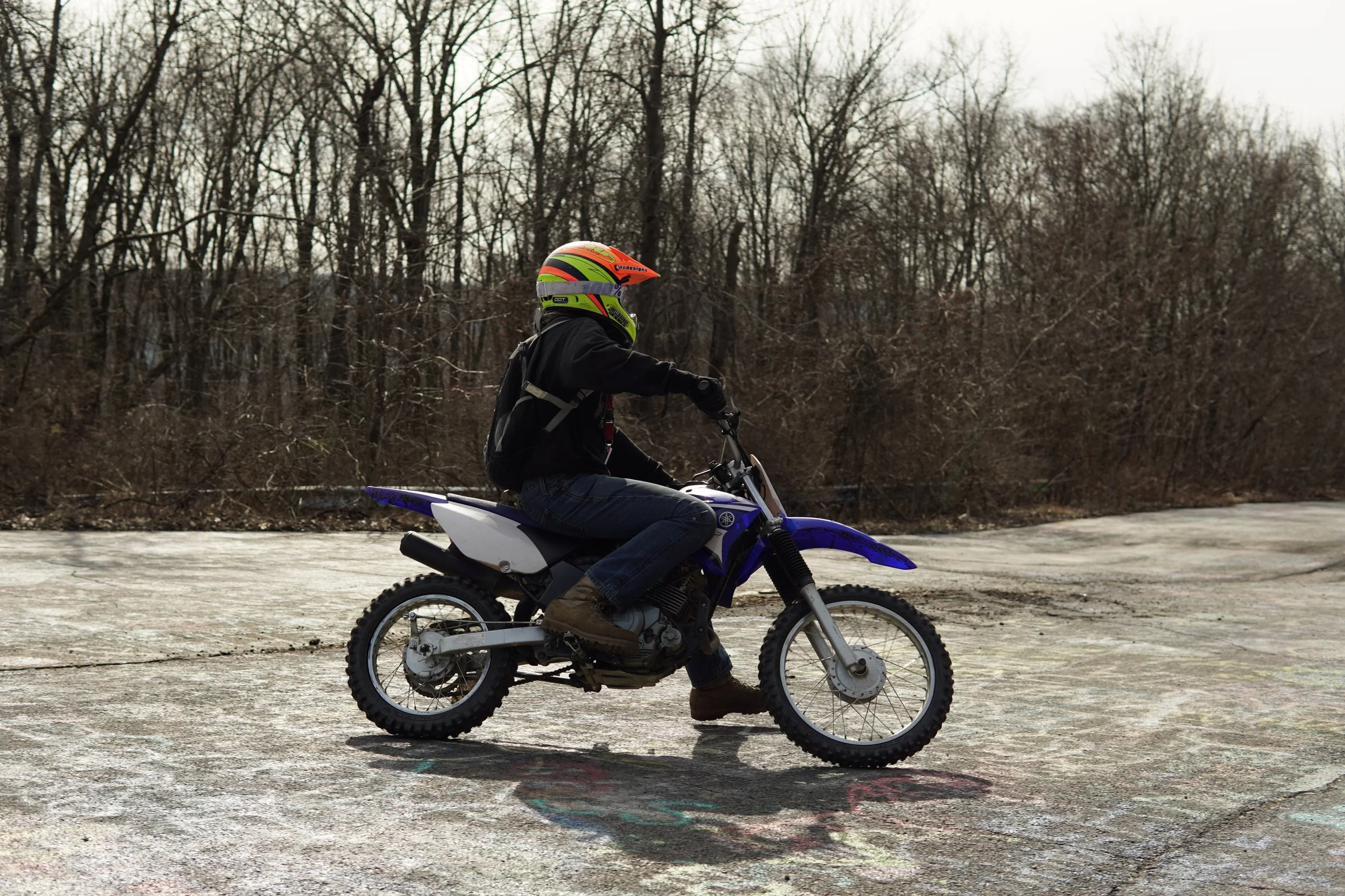 A person riding a blue and white dirt bike on an outdoor paved surface, wearing a black jacket, jeans, boots, and a colorful helmet, with leafless trees in the background.