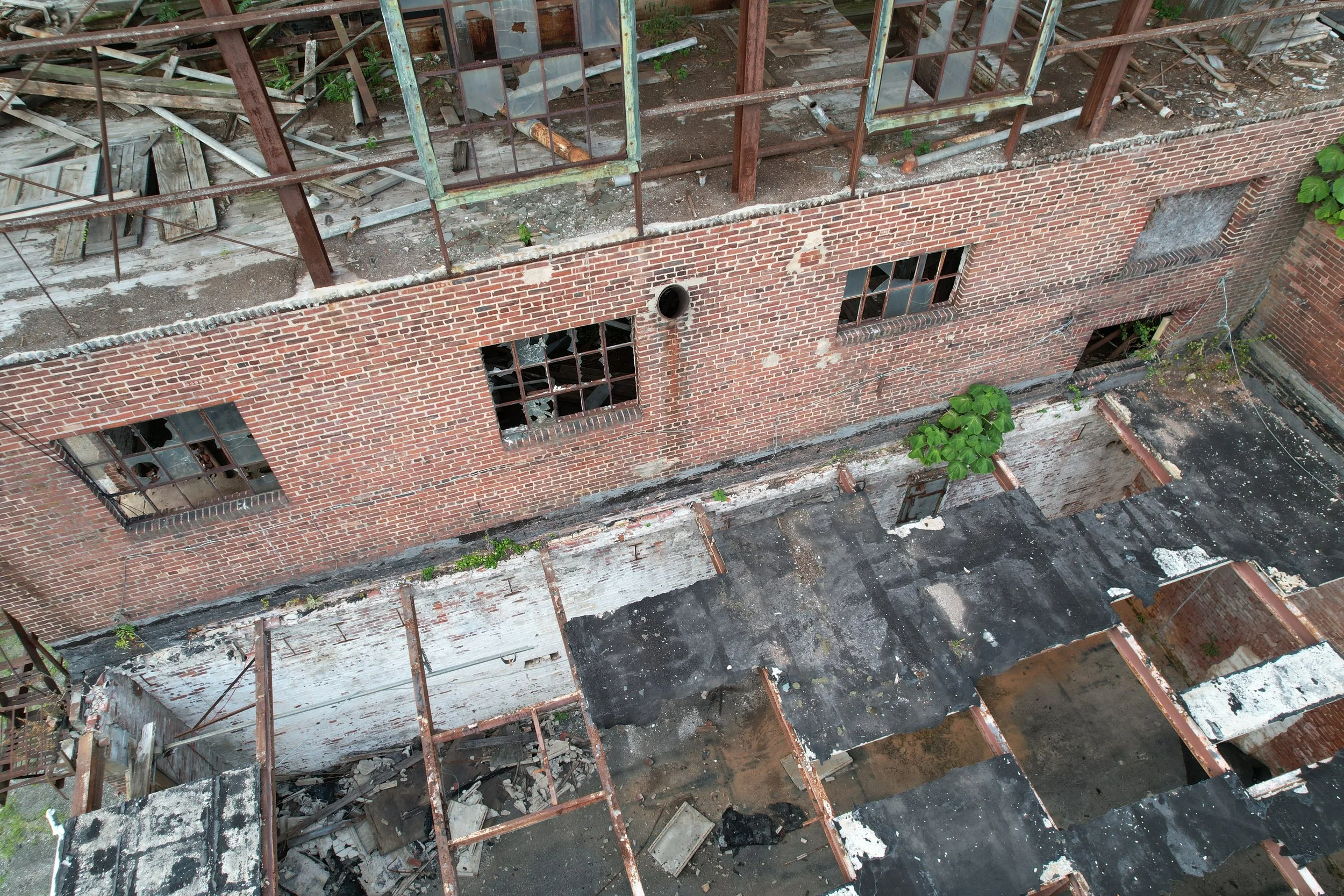 An aerial view of a dilapidated building with broken windows, exposed brick walls, and a collapsed roof with debris and overgrown plants.