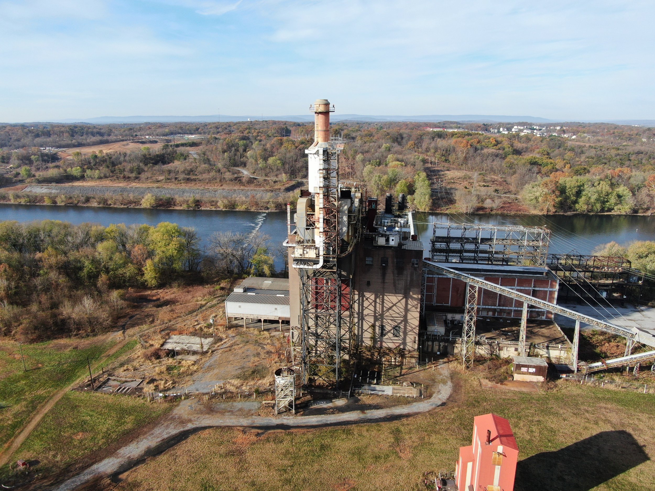 A power plant near a river with a forested landscape and distant mountains in the background, under a mostly clear blue sky.