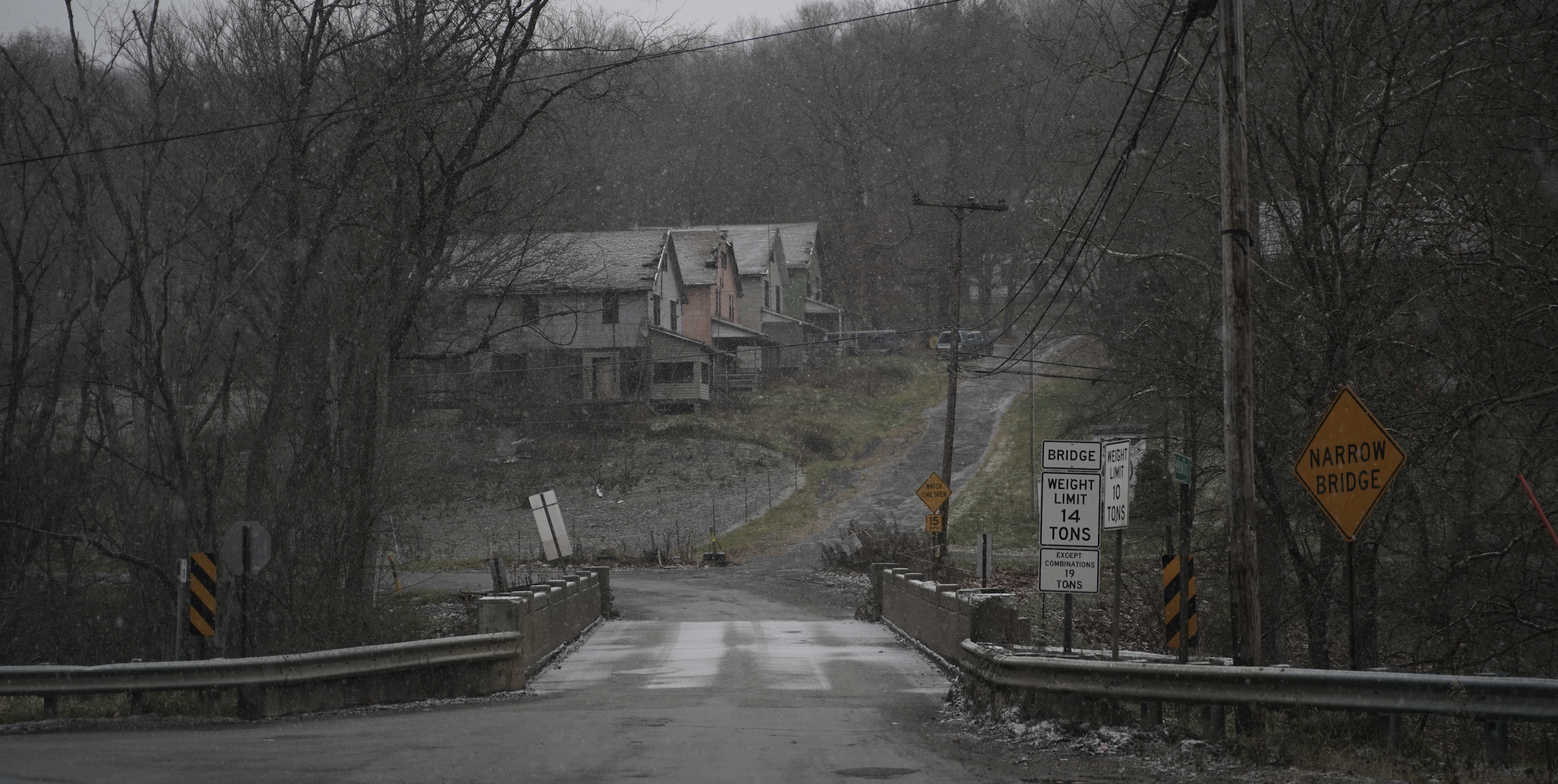 A small bridge with road signs indicating weight limits and narrow bridge warning, leading to residential houses on a hill in rainy weather, with leafless trees and power lines.