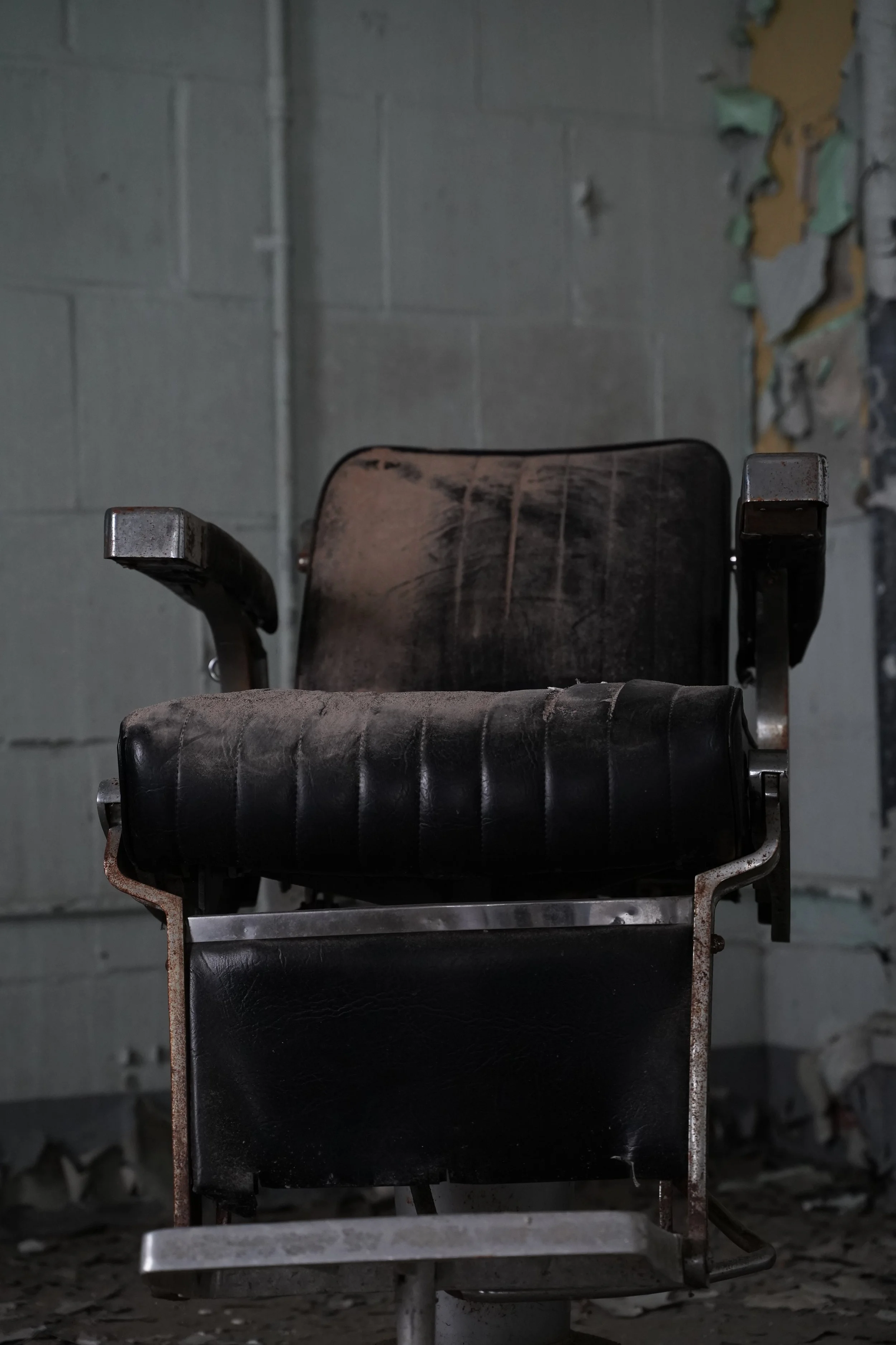 Old abandoned black leather barber's chair with rusted metal frame in an abandoned room with peeling paint and concrete block walls.