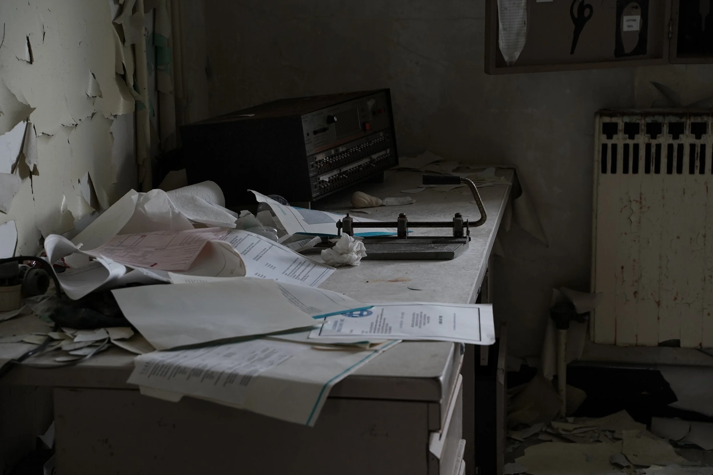 A cluttered, abandoned workspace with torn peeling paint on the wall, disorganized papers, a radio or electronic device, and a dirty, dusty surface in an old room.