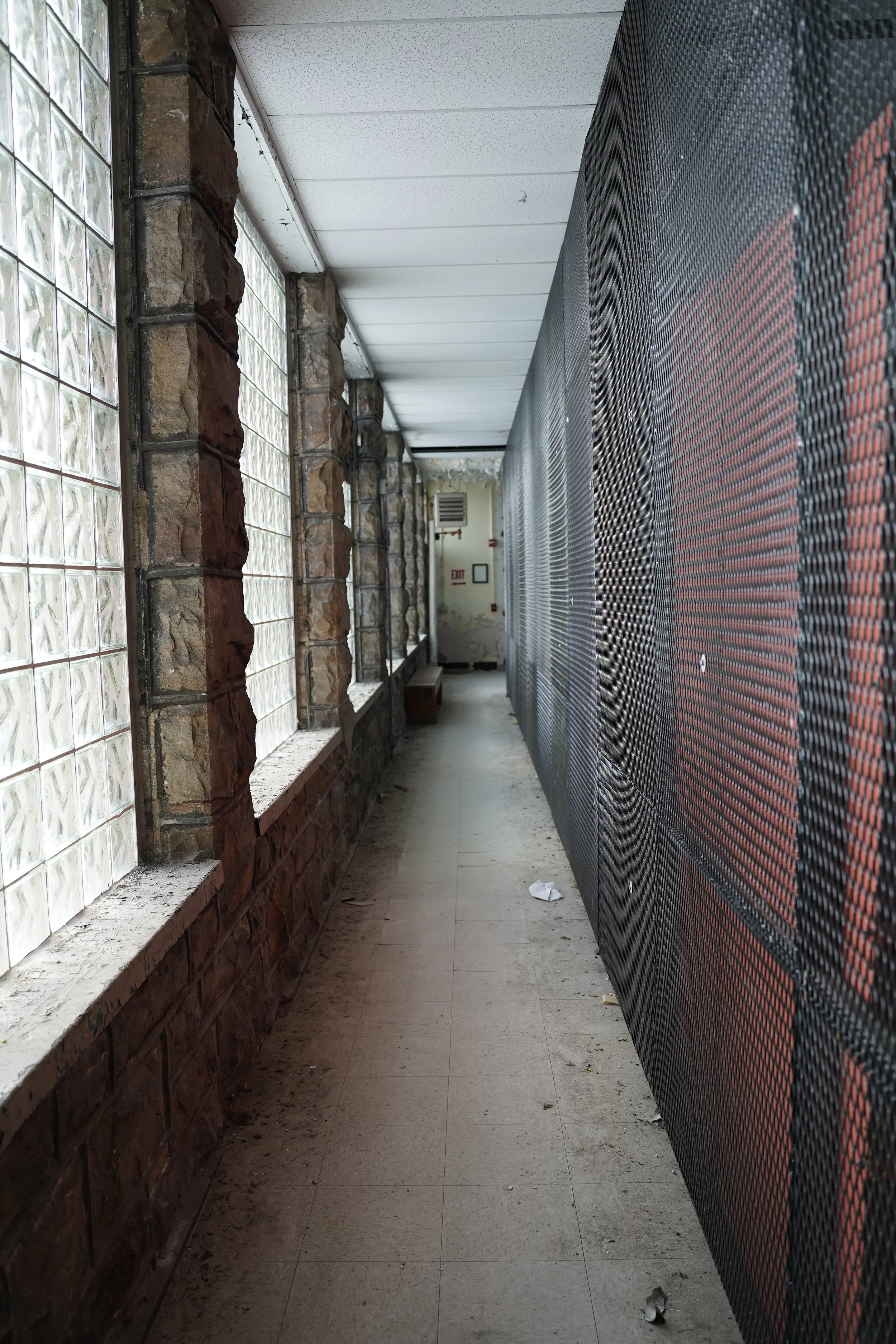 A corridor with a glass block wall on the left and a black perforated metal wall on the right. The floor is tiled, and there is a bench and an exit sign at the end of the corridor.