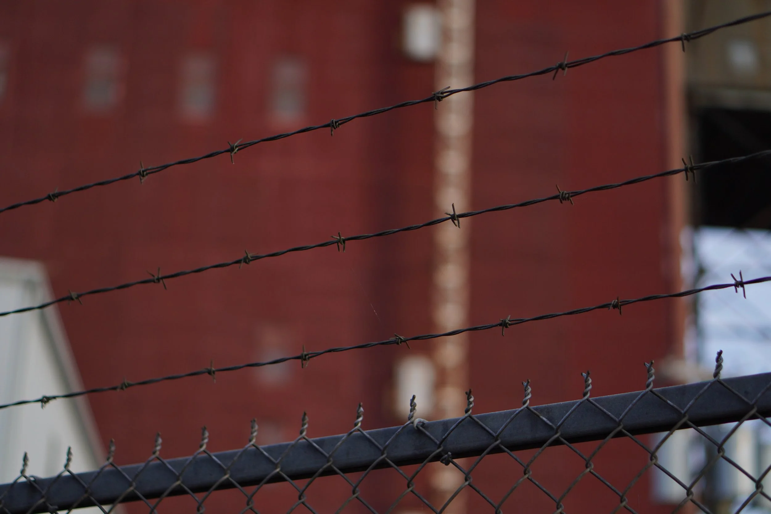 Close-up of black barbed wire fence with a red brick building in the background.