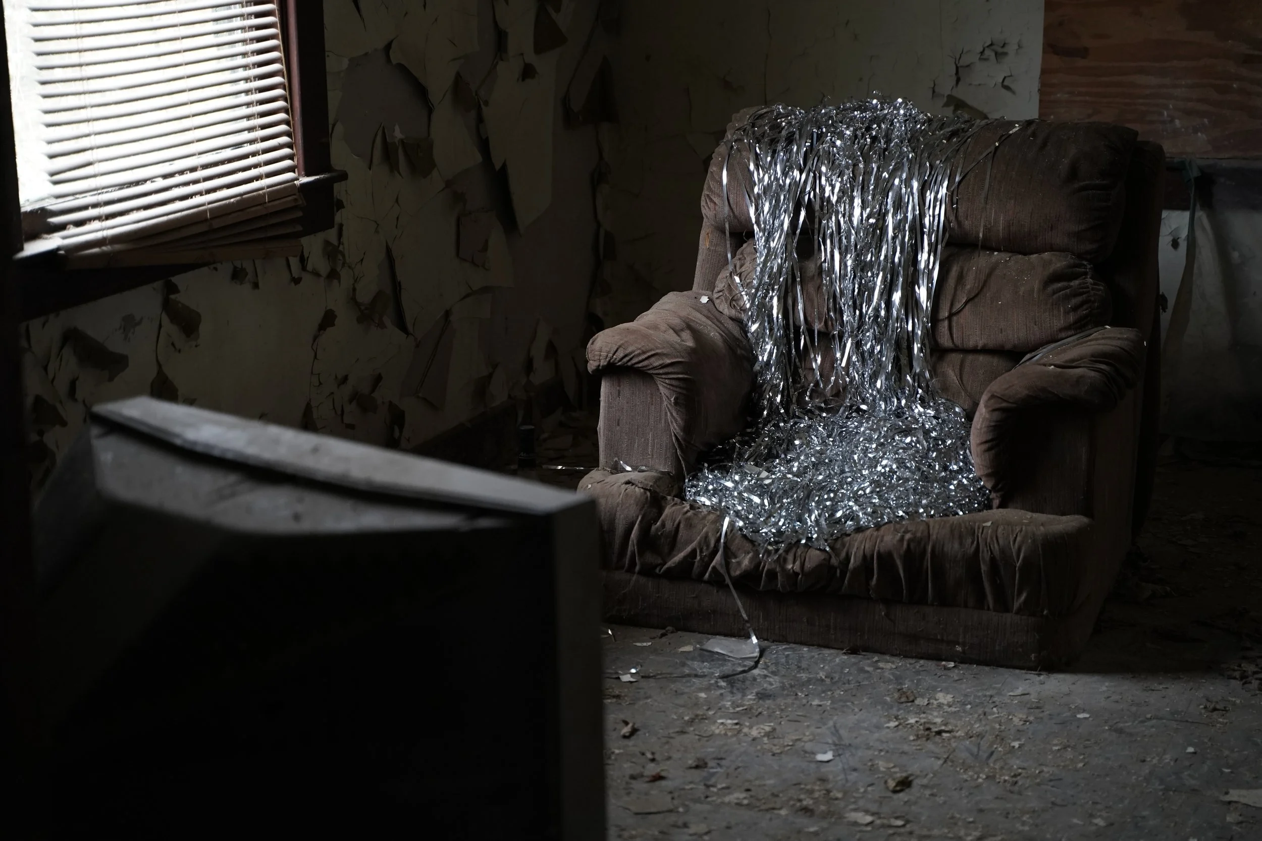 Old brown armchair in a dimly lit, abandoned room with peeling walls, covered with shiny silver tinsel.