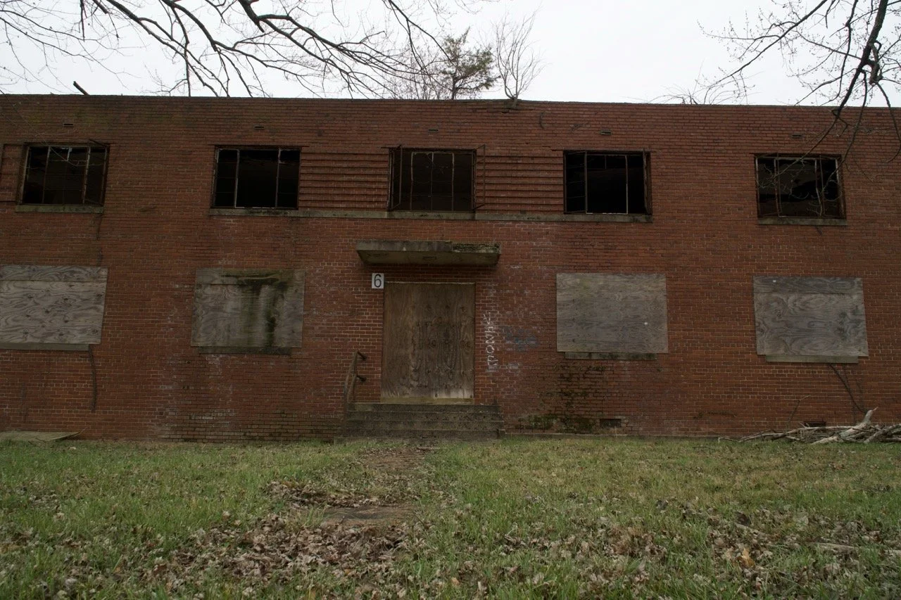 An abandoned two-story brick building with boarded-up windows and a boarded-up door, overgrown grass in front, and leafless tree branches overhead.