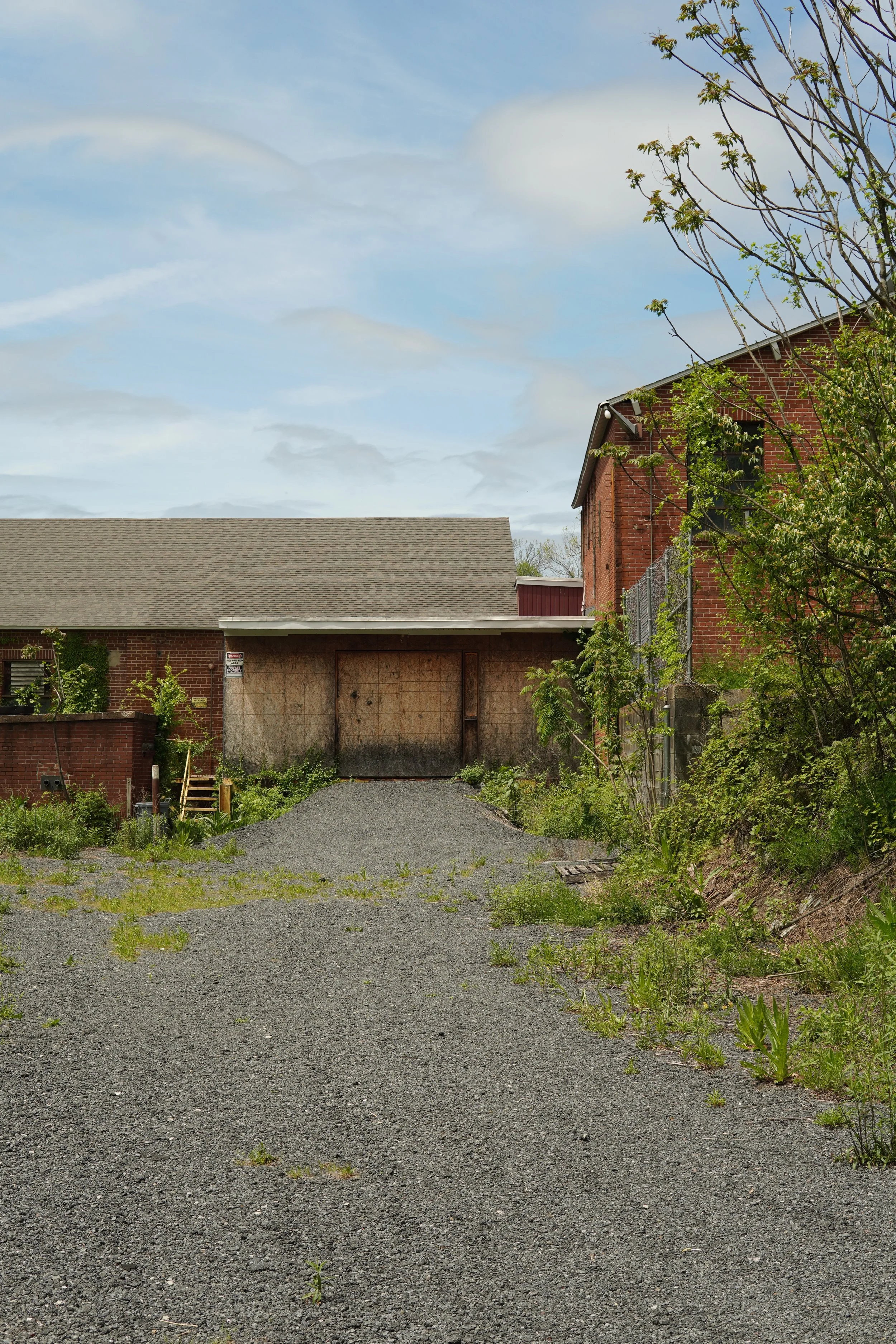 View of a partially boarded-up, red brick building with a large, flat gravel driveway in front. Overgrown weeds and small plants are scattered across the ground. A tree with budding leaves is on the right side of the image, and the sky is partly clou