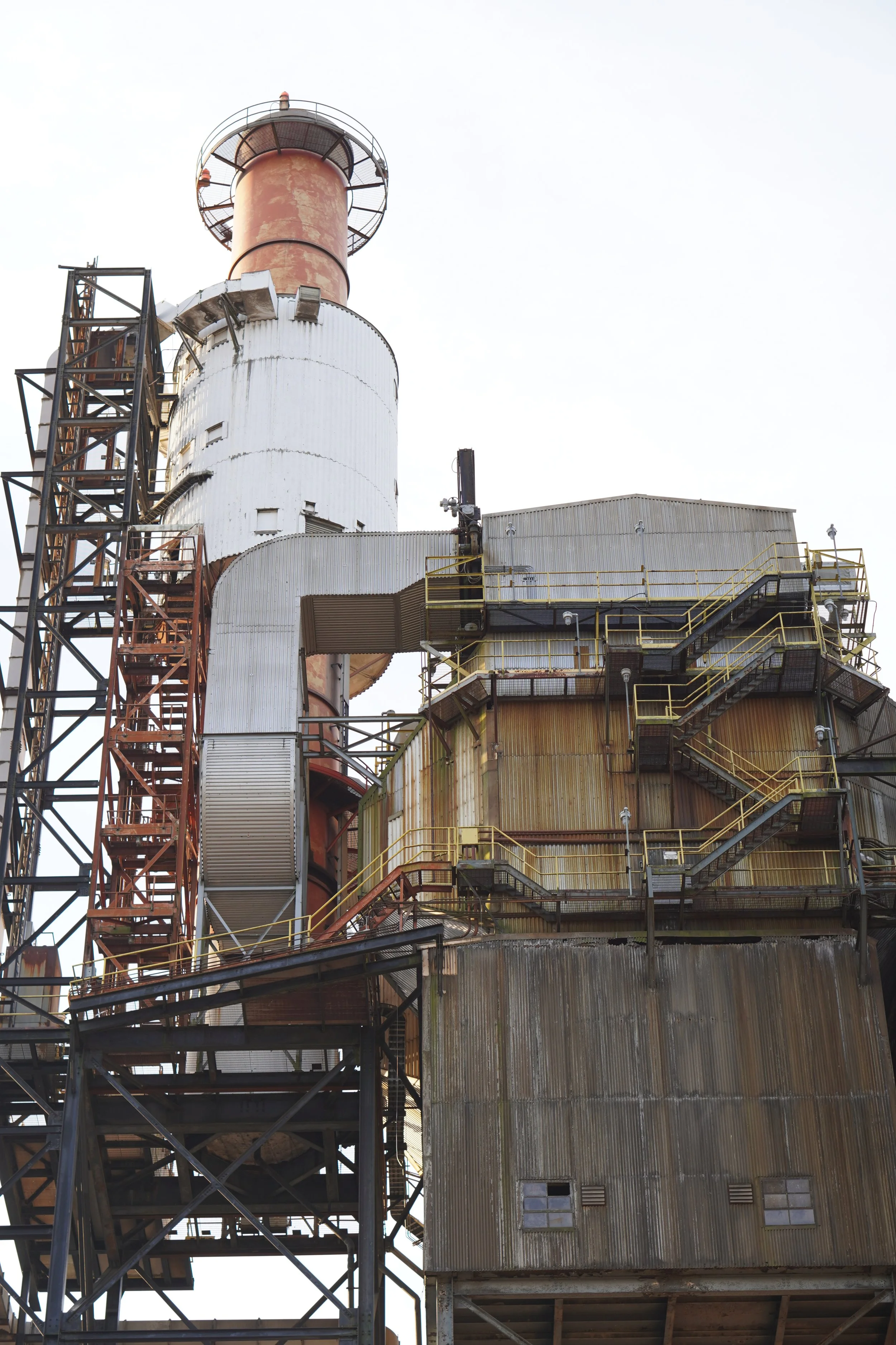 A tall industrial factory or refinery building with multiple levels of metal walkways, stairs, and pipes, showing signs of rust and aging.