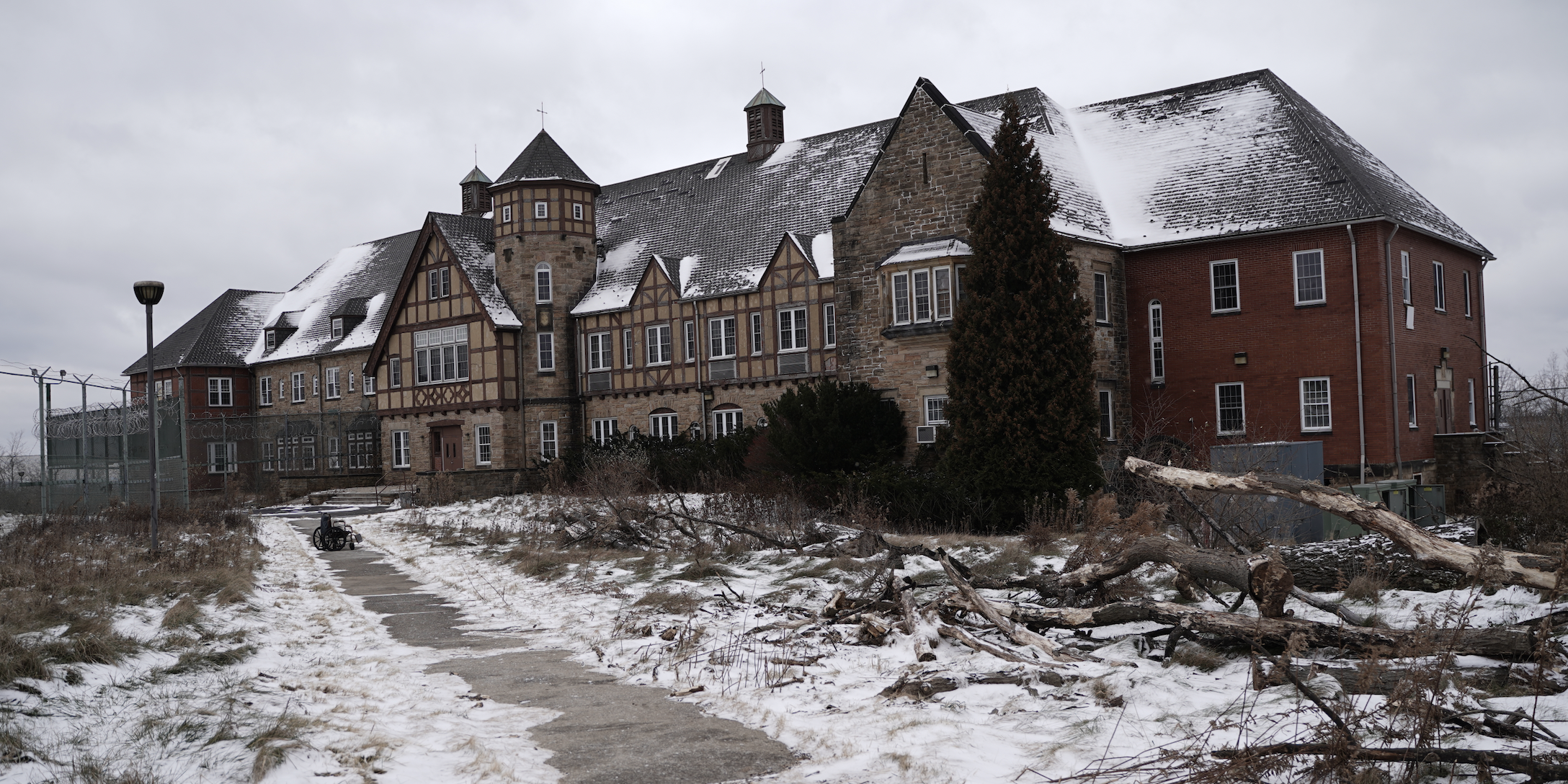 A large, old stone and brick building with a steep, snow-covered roof, several towers, and multiple windows. The scene is overcast, with patches of snow on the ground, fallen tree branches, a wheelchair on the pathway, and a tall tree in front of the building.
