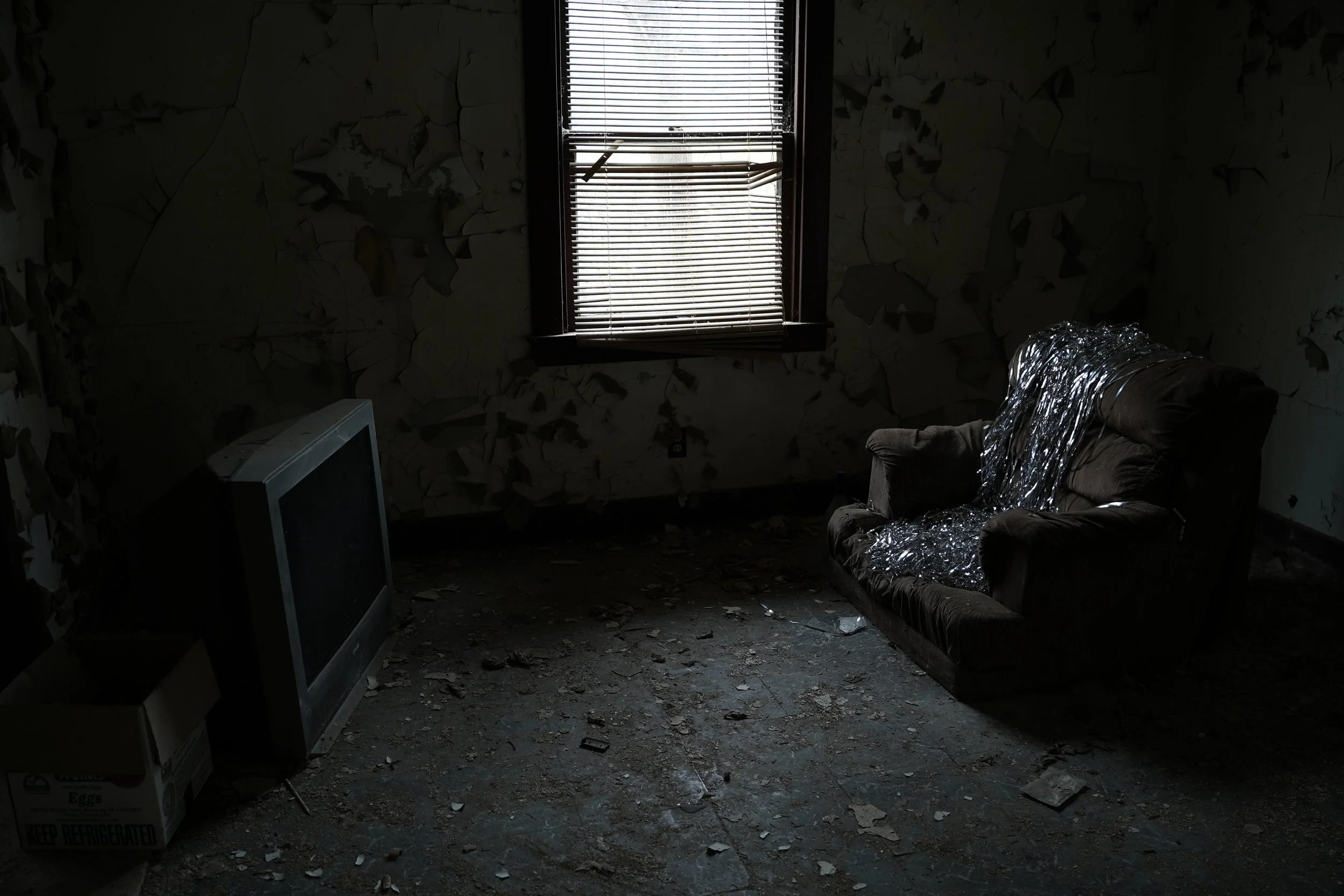 Dark room with peeling wallpaper, a window with broken blinds, a worn-out armchair with plastic wrap, a small discarded box, and an old television on the floor.