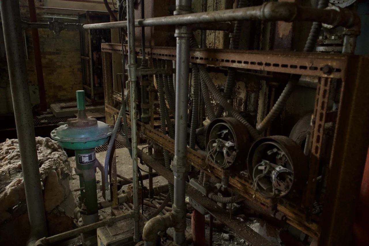 An industrial machine with exposed pipes, gears, and wiring in a dark, possibly abandoned factory or workshop.