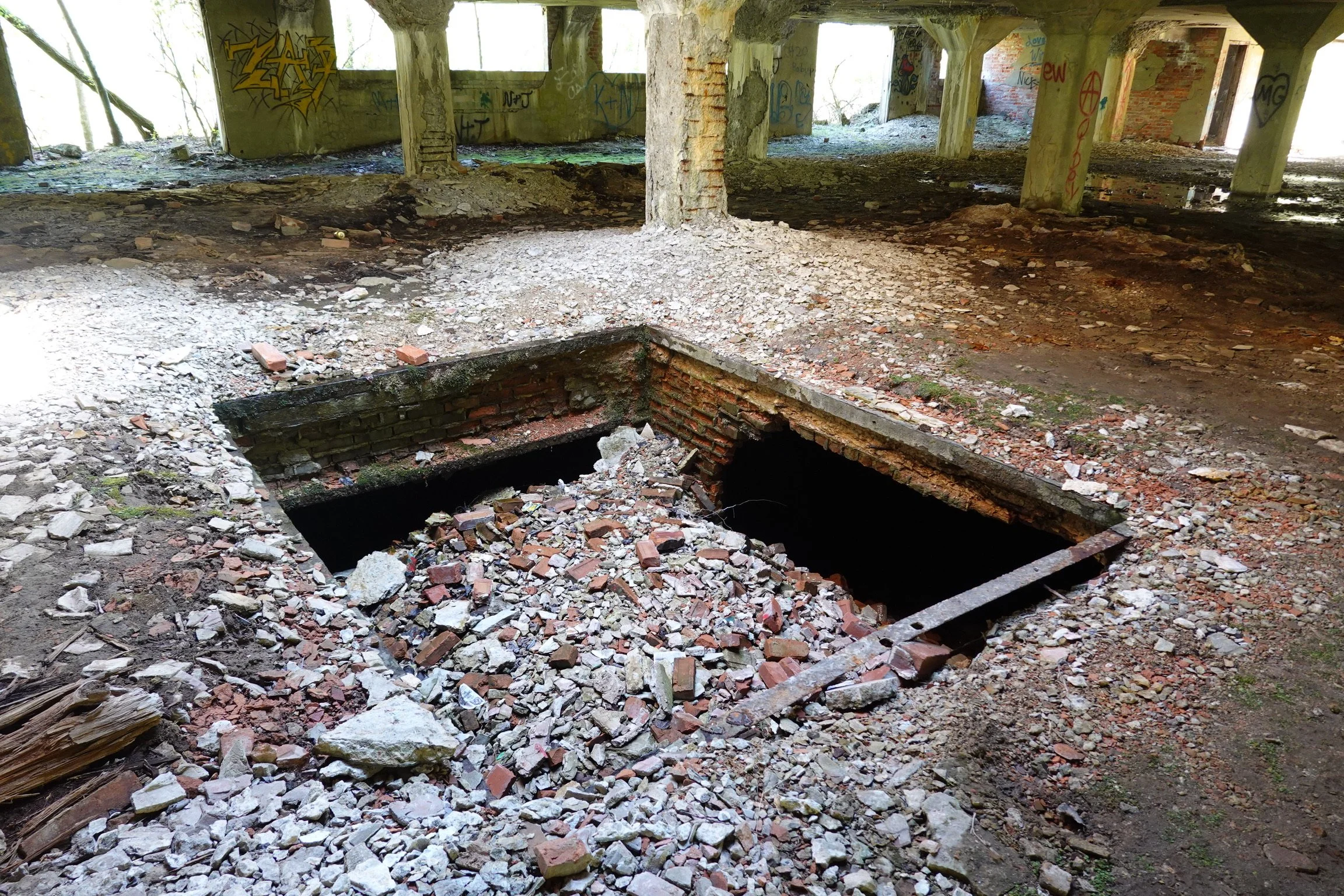 Interior of an abandoned building with open holes in the floor revealing below. The scene shows debris, bricks, and rubble scattered on the ground, with graffiti on the walls and supporting columns.