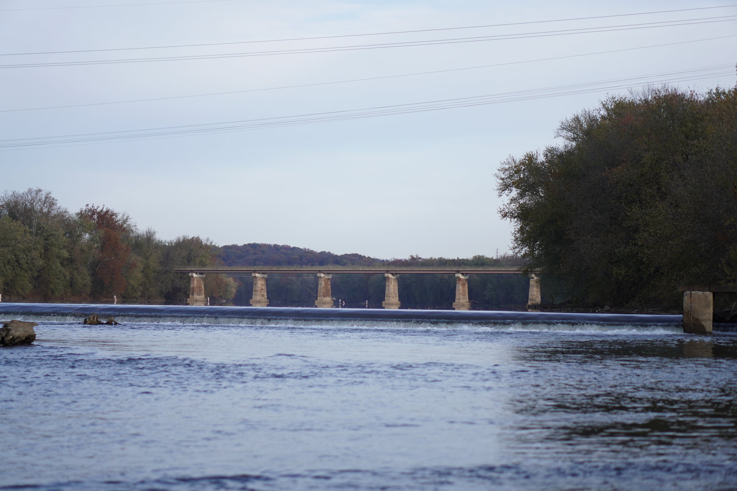 A river with calm water flow, a bridge over the river, trees on both sides with fall foliage, and a cloudy sky overhead.