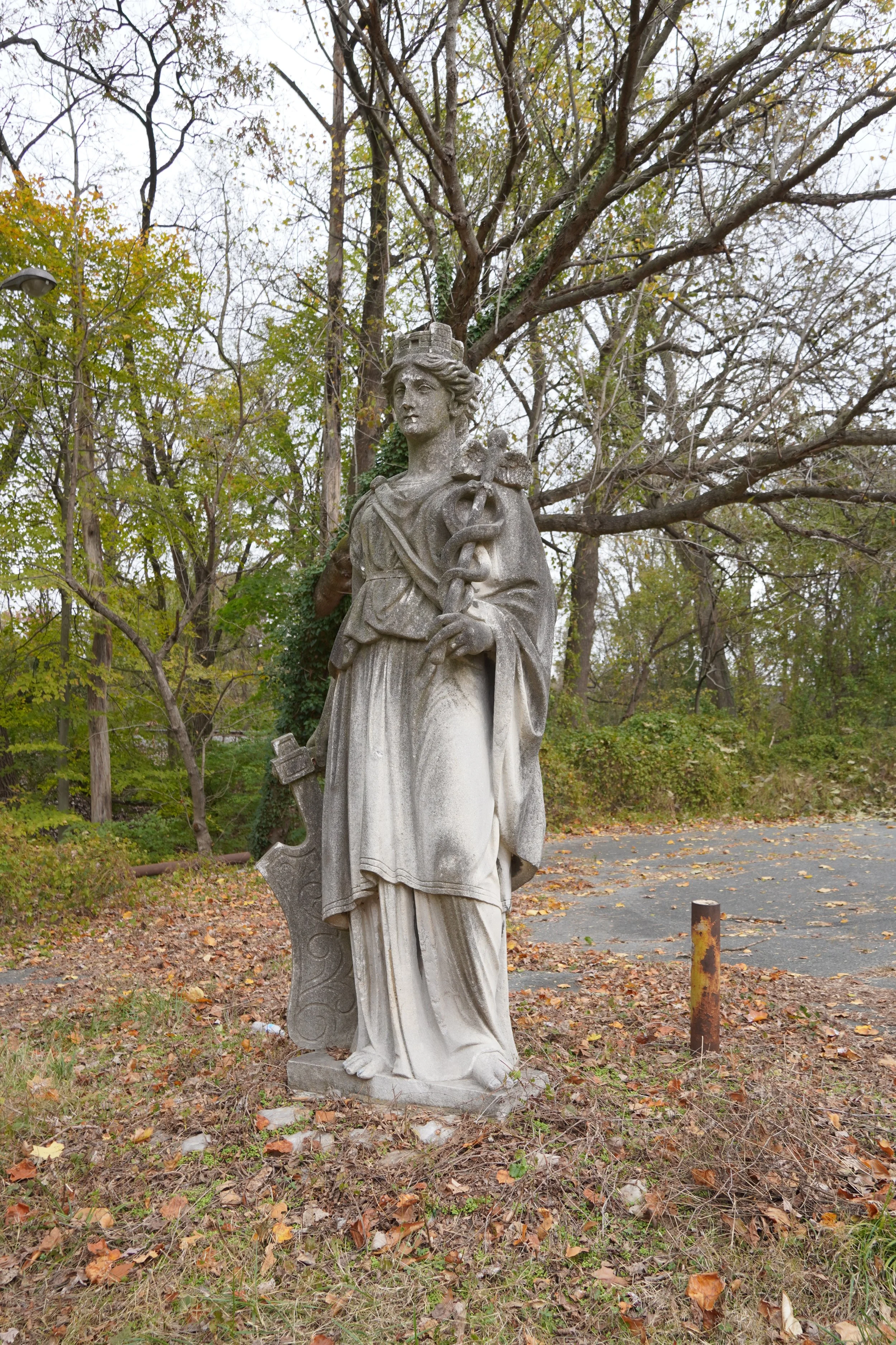 Statue of a woman with a shield and sword standing outdoors among trees with fall foliage.