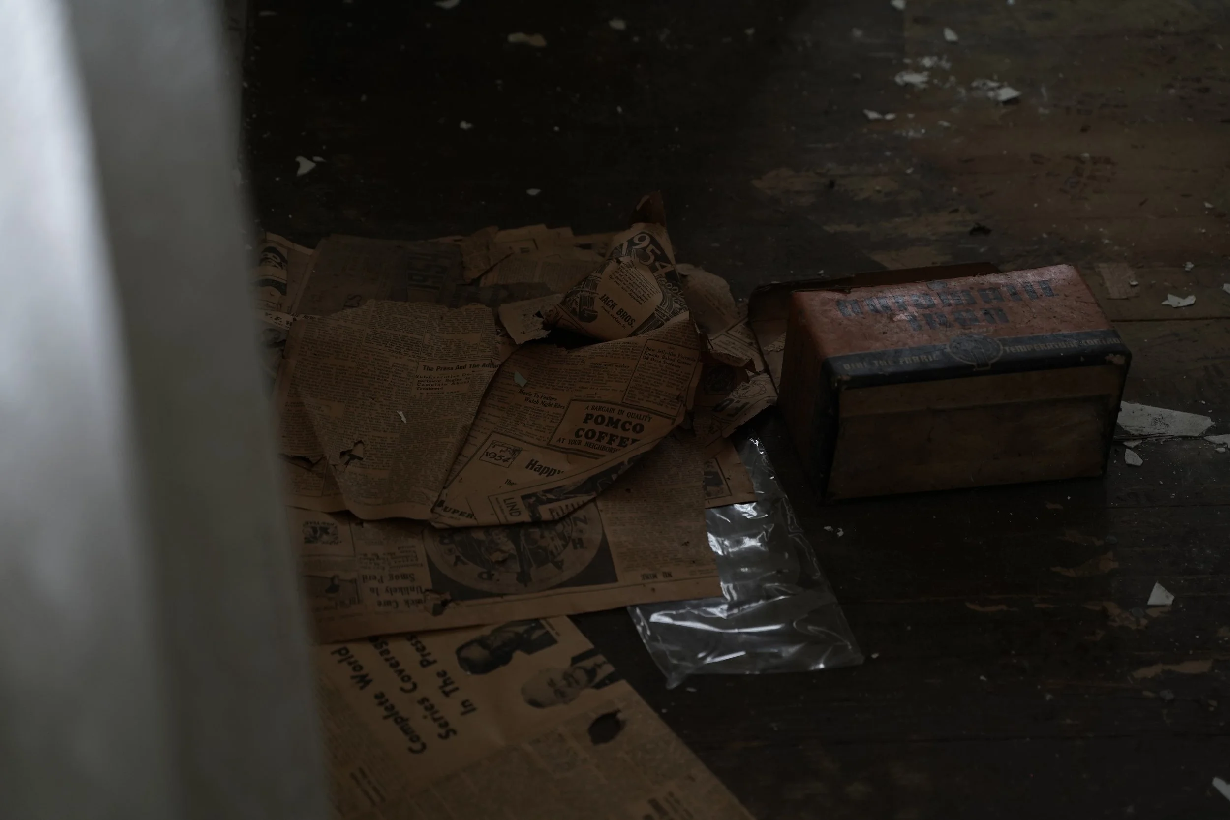 Dirty floor with scattered old newspapers, some torn, and a rusted metal box.